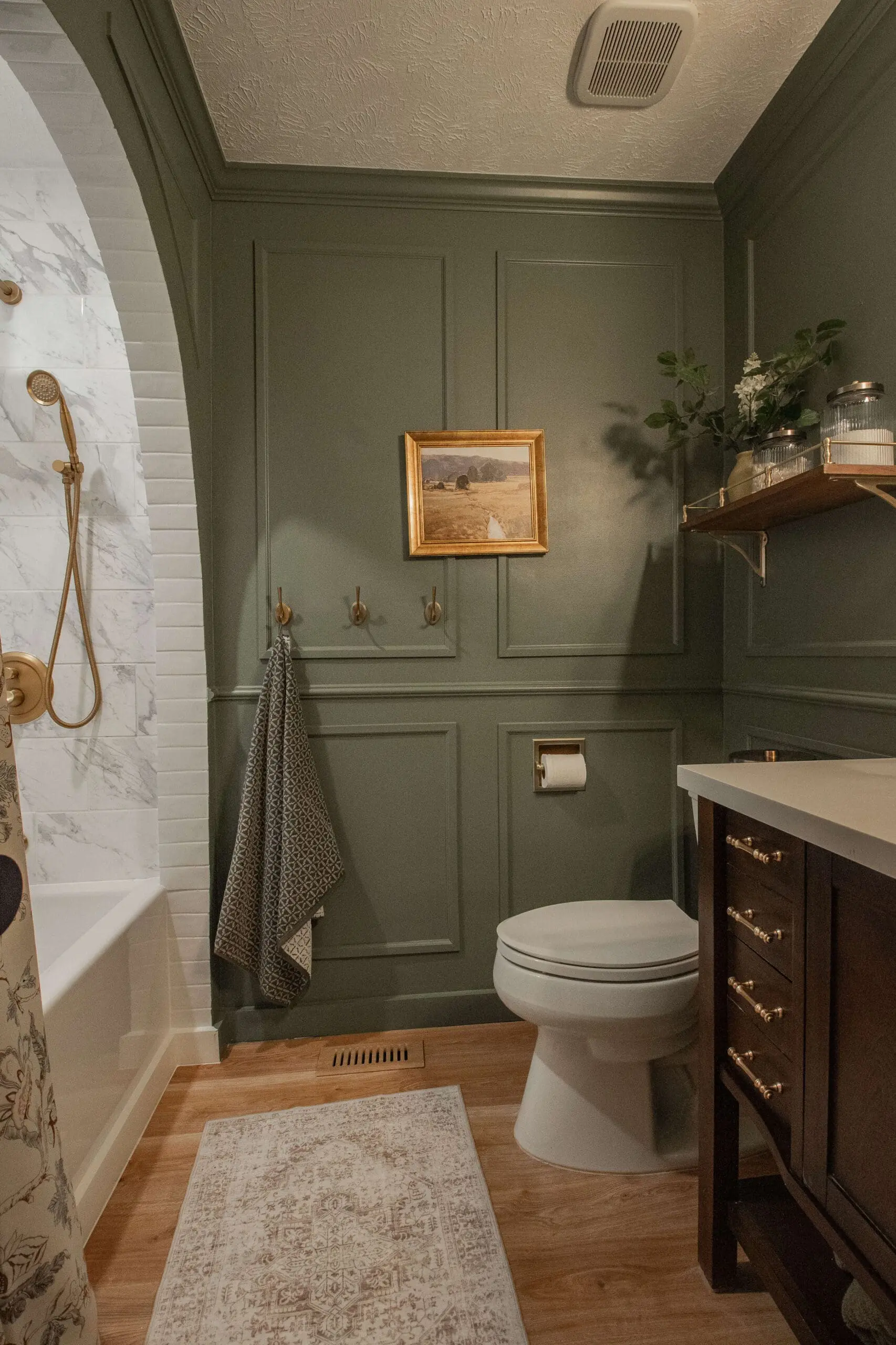 A cozy dark green bathroom with sage green paneled walls and a dark wood vanity with brass handles. The room includes a white toilet, a small gold-framed landscape painting, and three brass hooks holding a patterned gray towel. A wooden floating shelf adds a touch of warmth, styled with jars and fresh greenery.