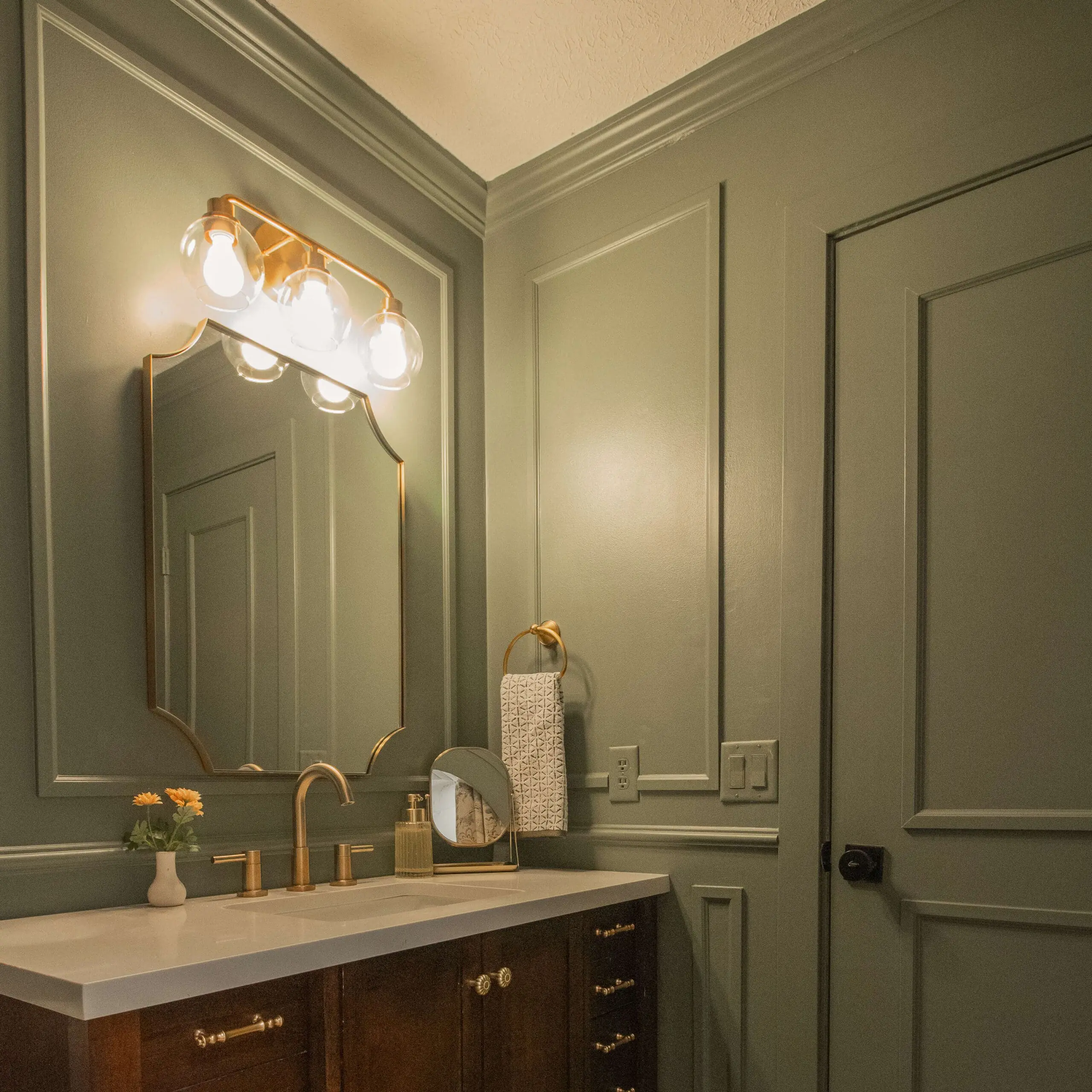 A sophisticated bathroom vanity with a dark wood base and gold hardware, paired with a crisp white countertop. The Sherwin Williams Dried Thyme walls feature DIY picture frame molding detailing, and a gold-framed mirror is illuminated by a trio of globe light fixtures. A gold towel ring holds a patterned hand towel, adding a refined touch.