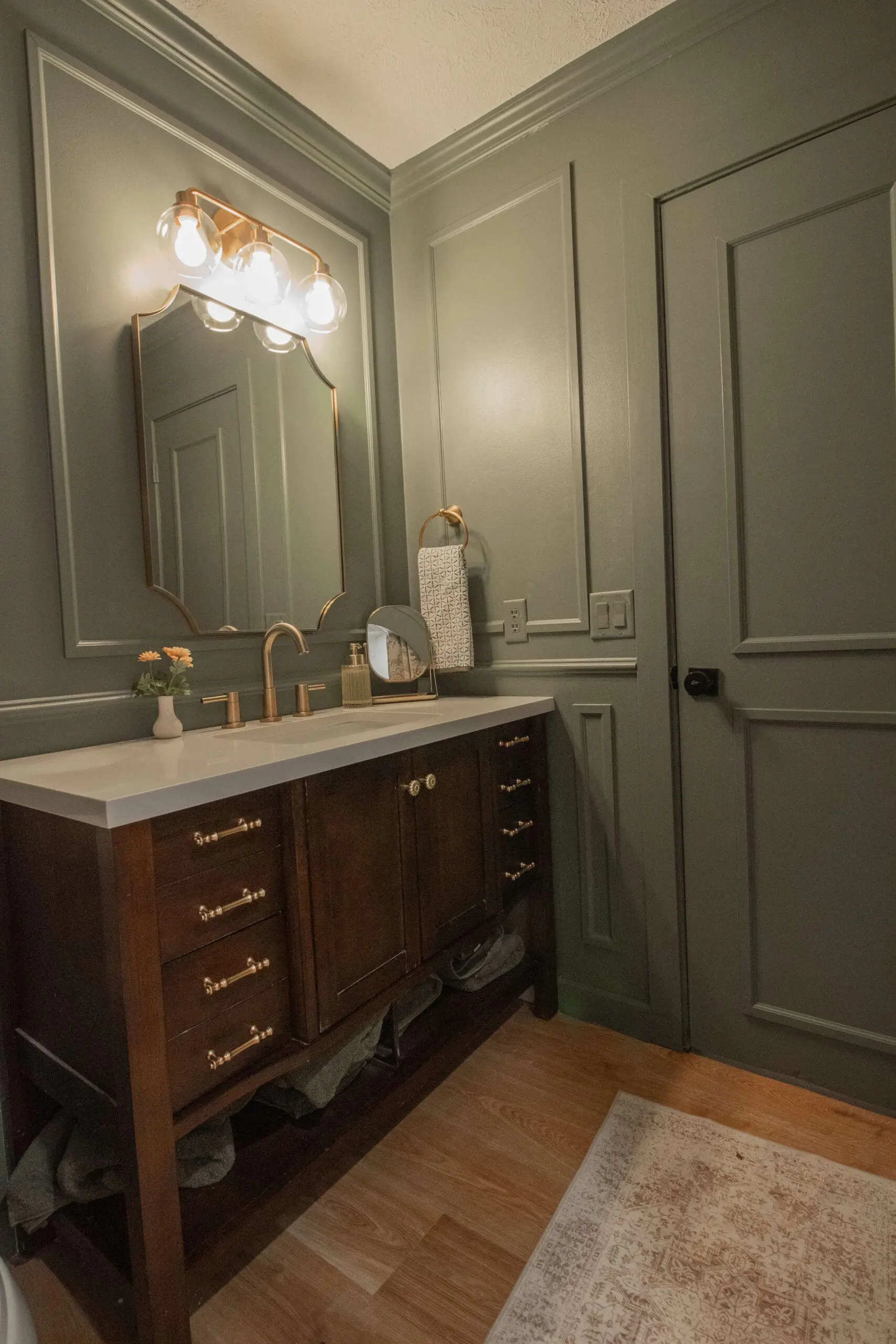 A charming bathroom with sage green paneled walls, a white countertop, and a dark wood vanity with brass knobs and handles. A gold-rimmed mirror and gold light fixture add elegance above the sink area. The door is painted to match the walls, creating a cohesive and calming aesthetic.