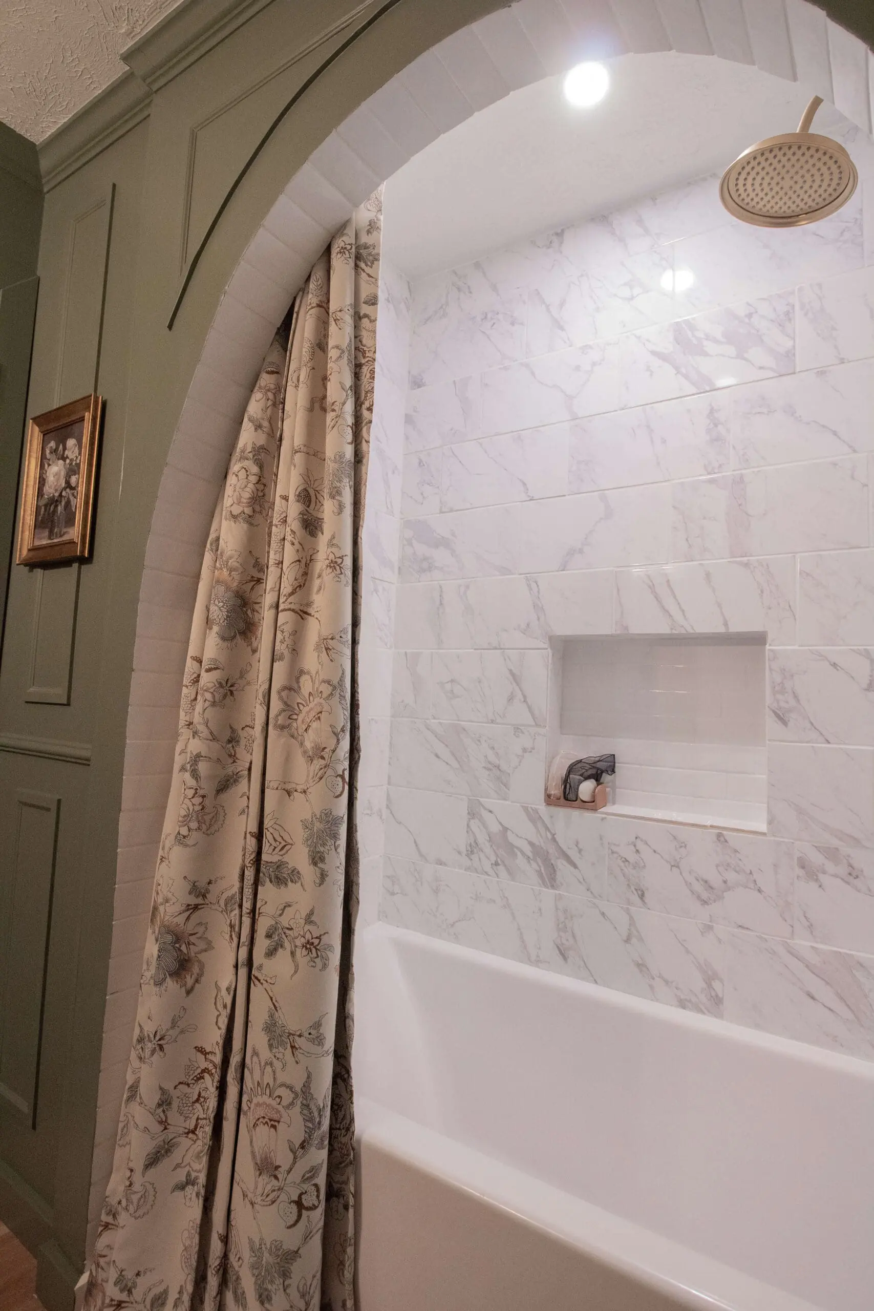 A chic bathroom showcasing a tub surrounded by marble tiles, an arched frame with white subway tiles, and brass fixtures. A floral curtain and a built-in wall niche add character, while the green wainscoting adds a refined contrast.