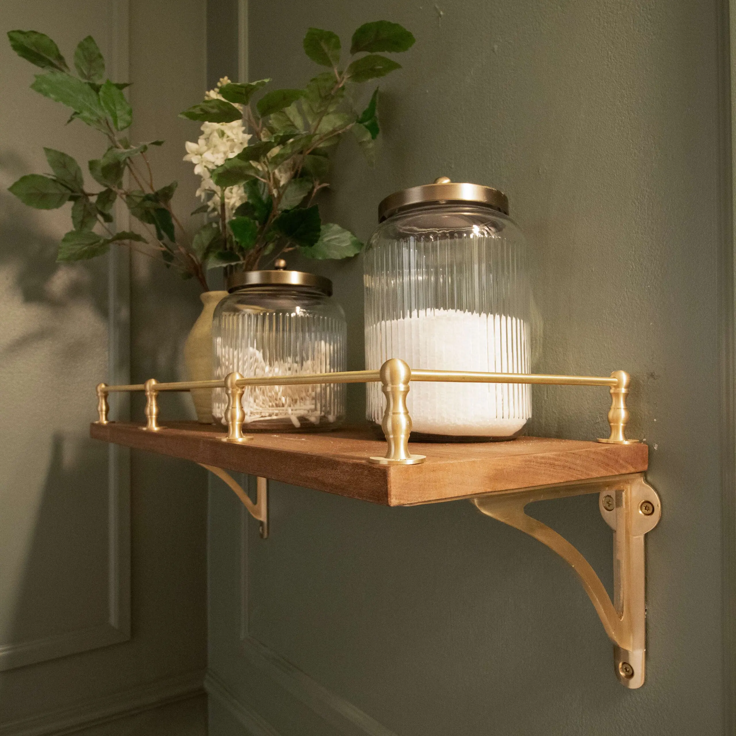 Angled view of a wooden shelf featuring a brass rail, holding two glass jars and a vase with flowers. The gold brackets stand out against the deep green paneled wall, creating a luxurious, modern aesthetic.