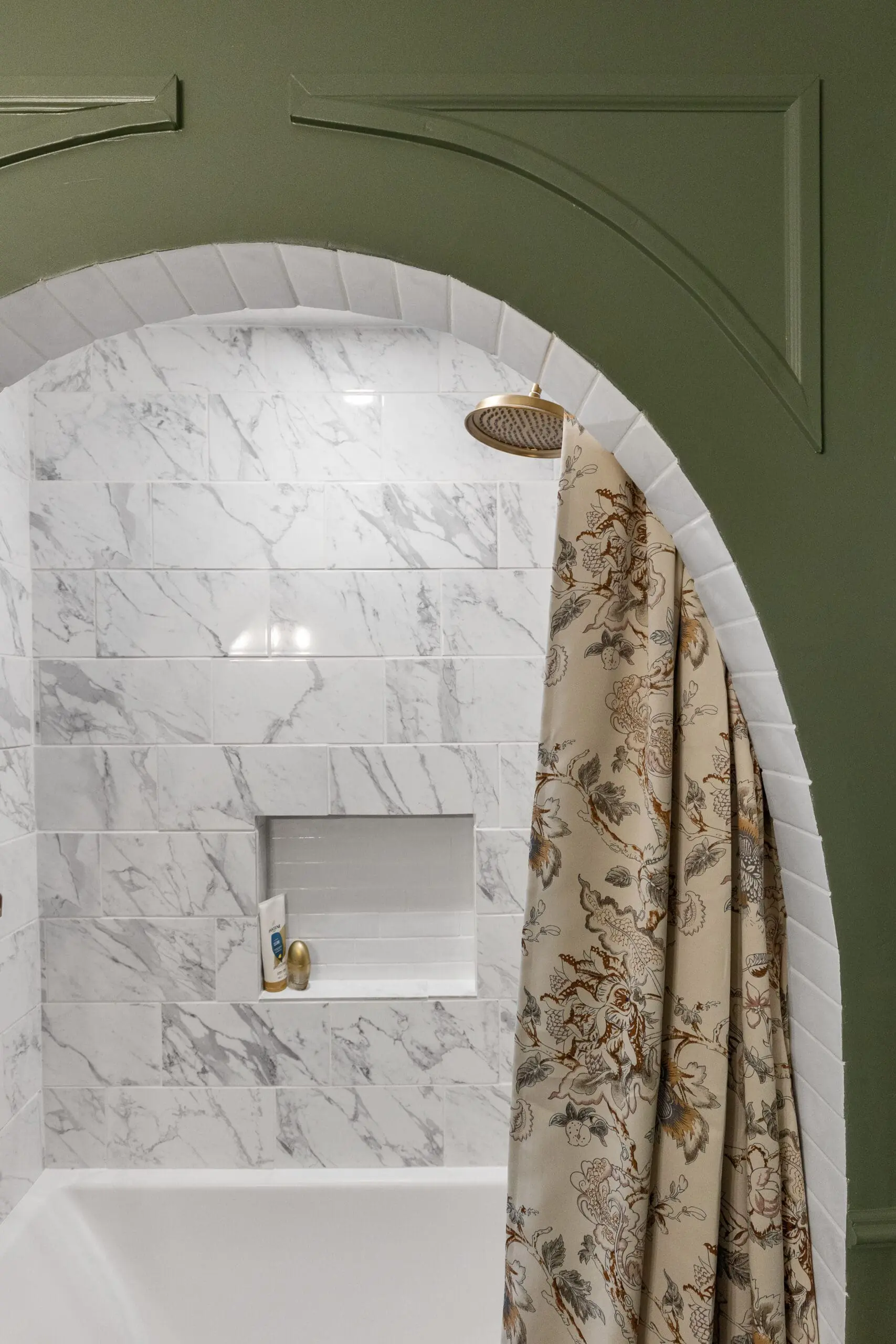 Arched shower with deep green trim, floral shower curtain, and marble wall tile surrounding a built-in shower niche and brass fixtures.