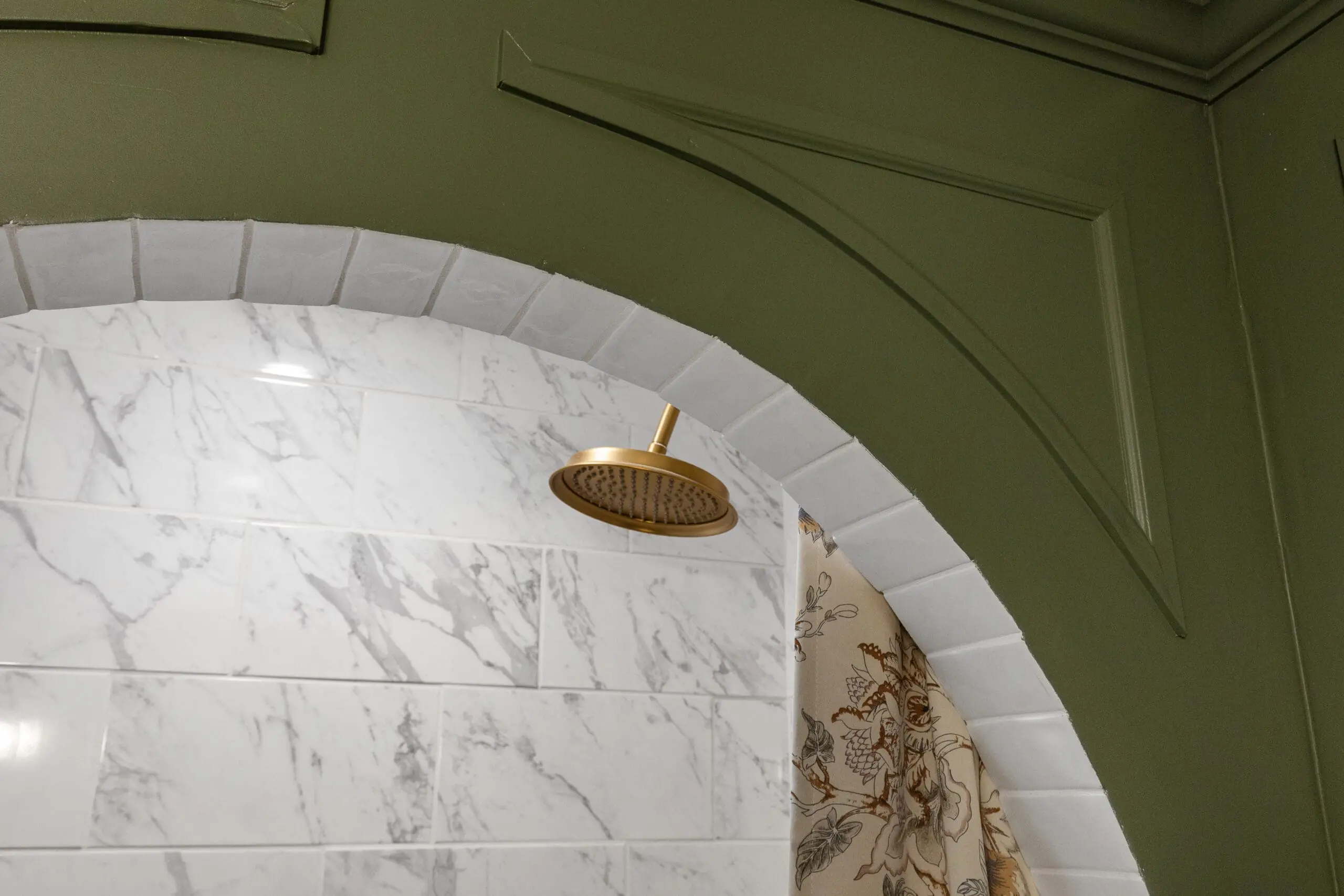 Brass rain showerhead installed in a marble-tiled shower with a white subway-tiled arch and green-painted molding detail.