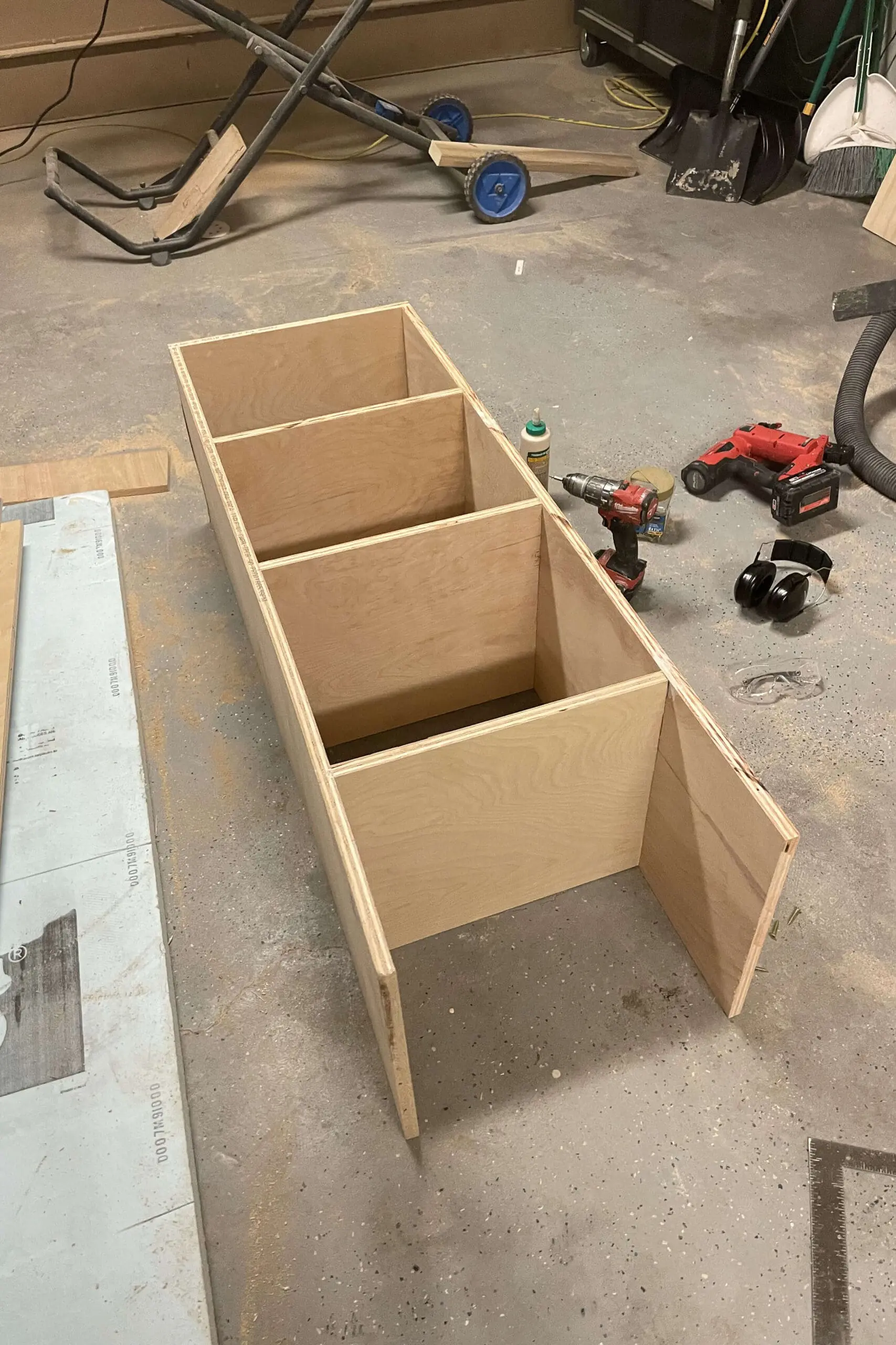 Unfinished wooden cabinet frame lying on a workshop floor, with compartments and dividers taking shape. Tools, including a drill, nail gun, safety goggles, and wood glue, are scattered nearby, indicating an ongoing DIY project.