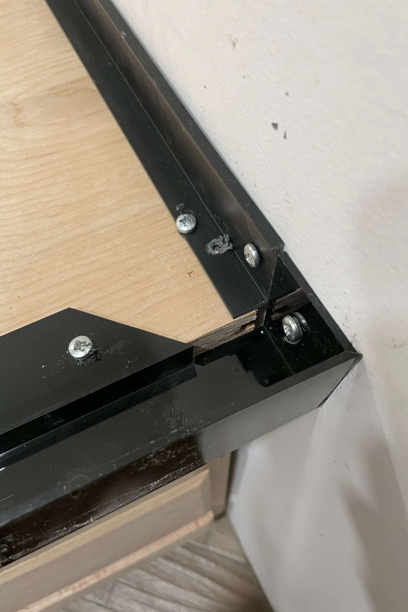 Close-up of black plastic pieces forming a corner frame on the wooden countertop base, secured with screws. The corner joint creates a mold for a concrete countertop, with the white wall in the background
