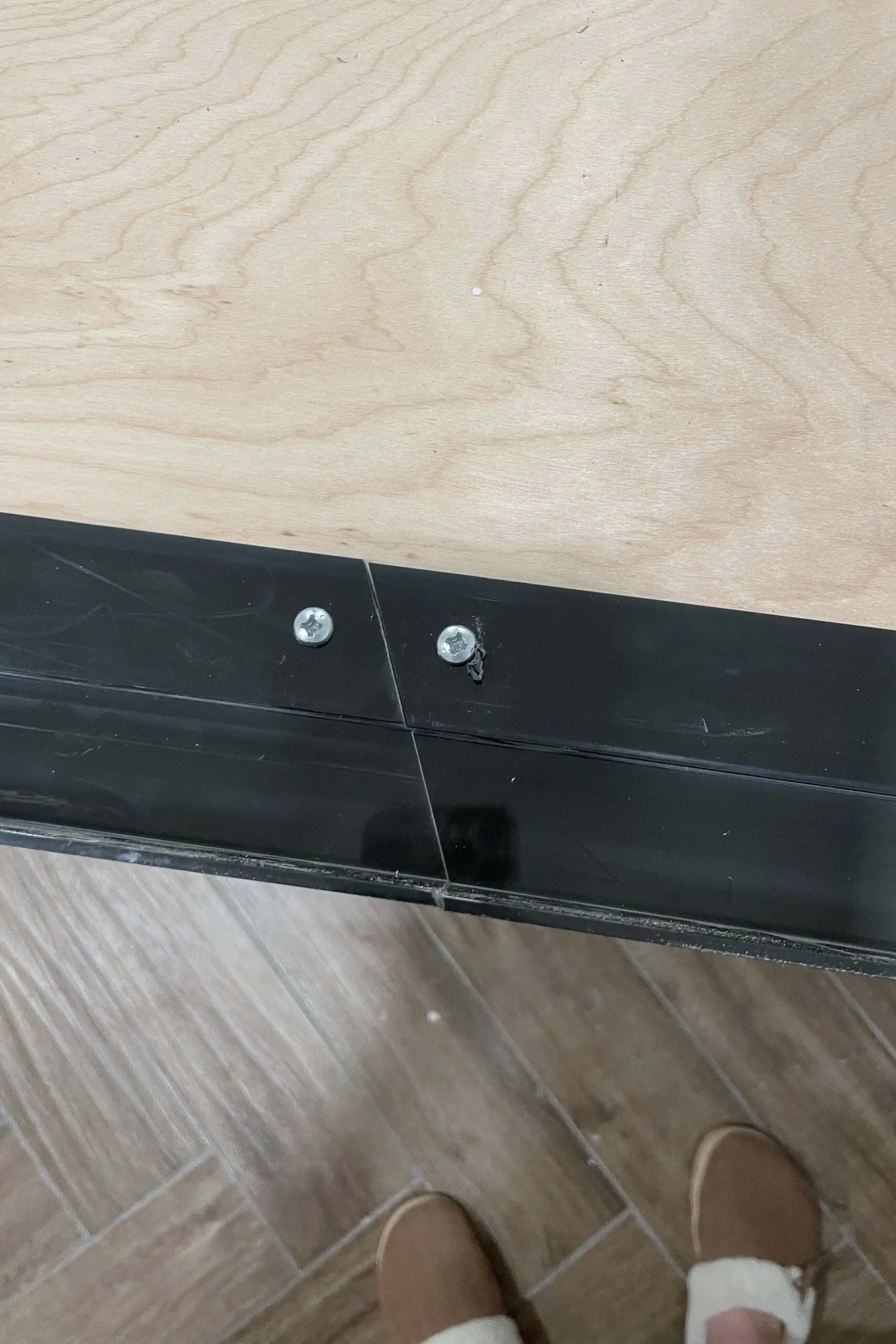 Detailed view of black plastic concrete forms secured to the wooden countertop base with screws. The pieces meet at a seam, creating a form for a concrete countertop, with natural wood grain visible on the surface.