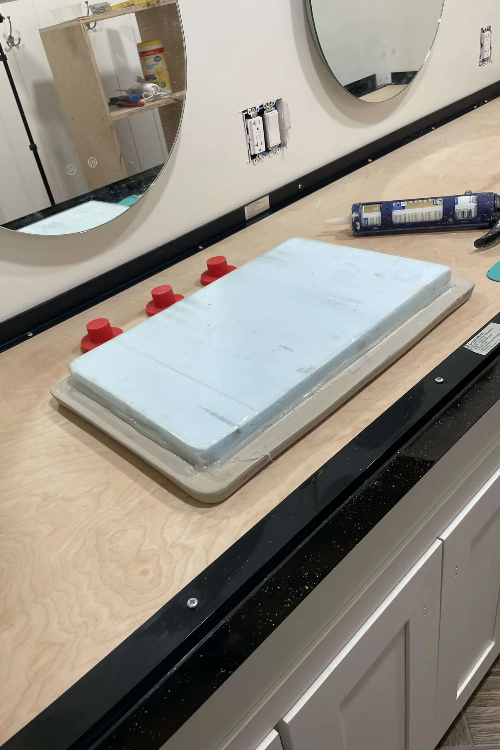 Bathroom vanity under construction with a wooden base surrounded by black plastic frame pieces to create a mold for a concrete countertop. On the wooden surface are a wrapped foam block and plastic knockouts, while oval mirrors and an exposed electrical outlet are mounted on the wall.