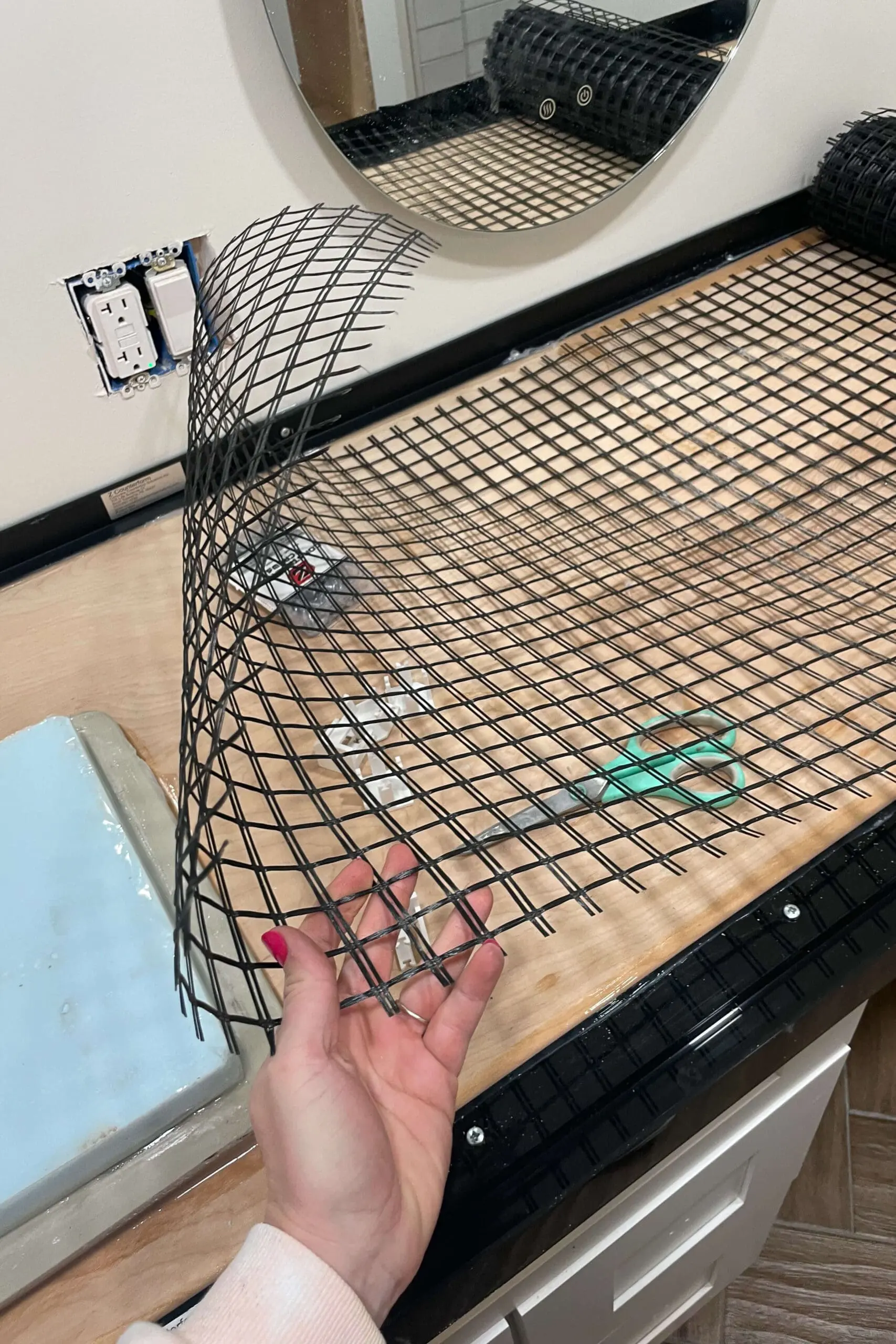 Hand holding a flexible black plastic mesh over a wooden base with black plastic framing, preparing the formwork for a concrete countertop. A green-handled pair of scissors rests on the wooden surface, and an oval mirror is mounted on the white wall in the background.