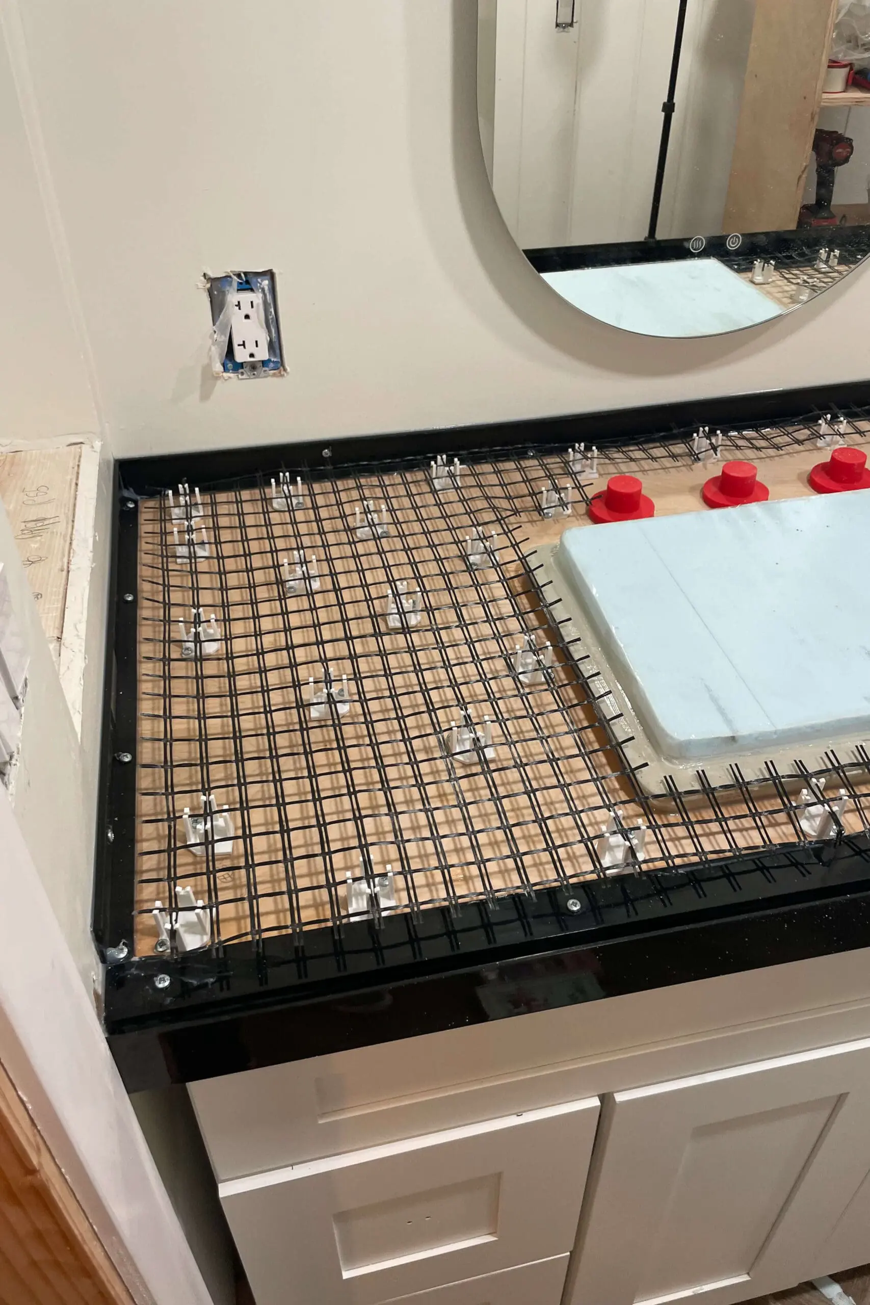 Bathroom vanity under construction with a black plastic mesh secured over a wooden base surrounded by black plastic framing. The mesh is supported by small white spacers to create reinforcement for the concrete countertop. Red knockouts and a foam block are placed on the surface, with an oval mirror and an exposed electrical outlet visible on the white wall.