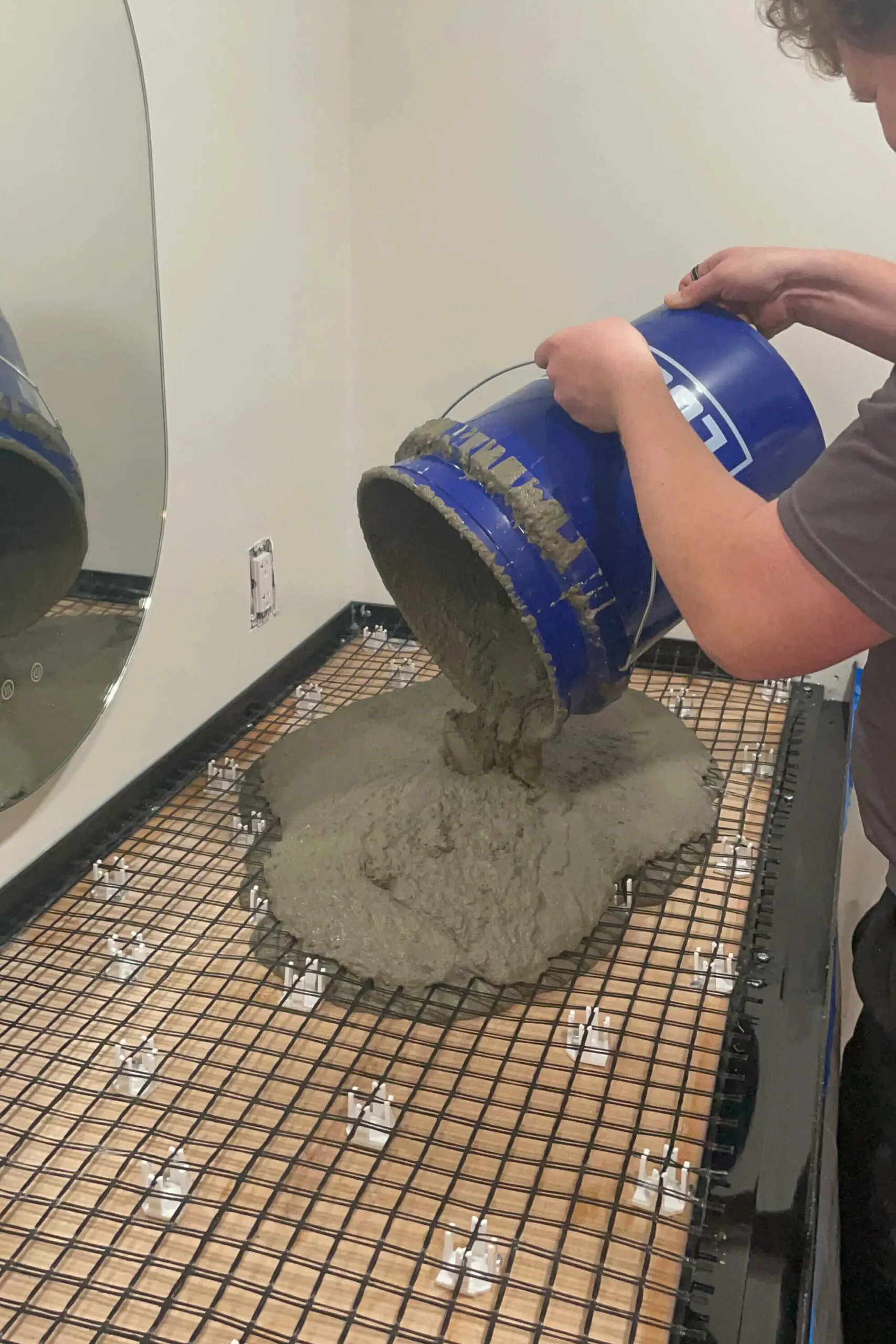 A person pouring wet concrete from a blue bucket onto a black plastic mesh grid over a wooden base, forming the foundation of a bathroom countertop. The process is part of a concrete countertop installation project.