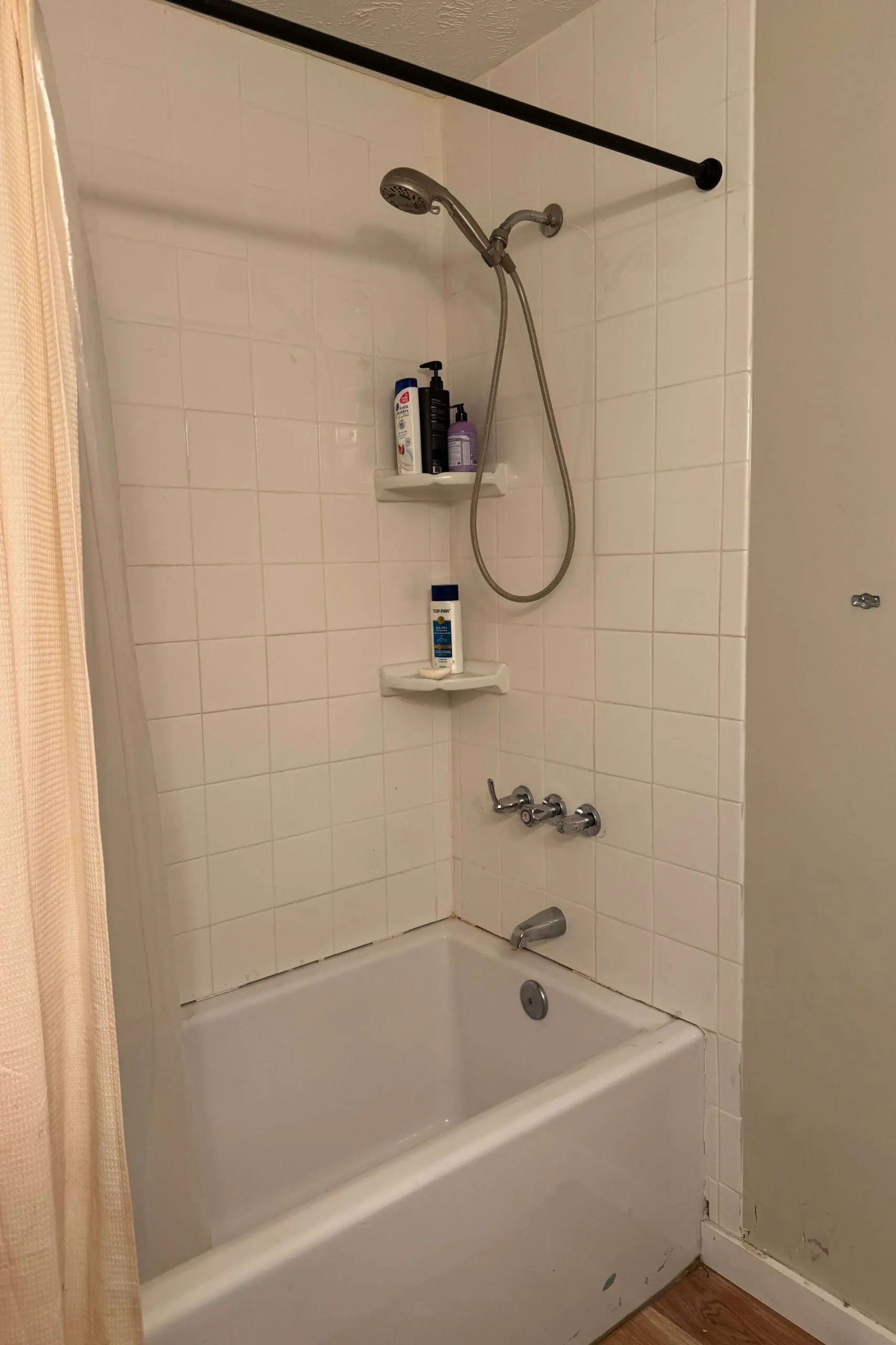 A standard white bathtub with white tiled walls and two small corner shelves holding toiletries. A black shower curtain rod frames the space, with a handheld showerhead attached. The surrounding area shows some wear, with visible seams where the tiles meet the wall.