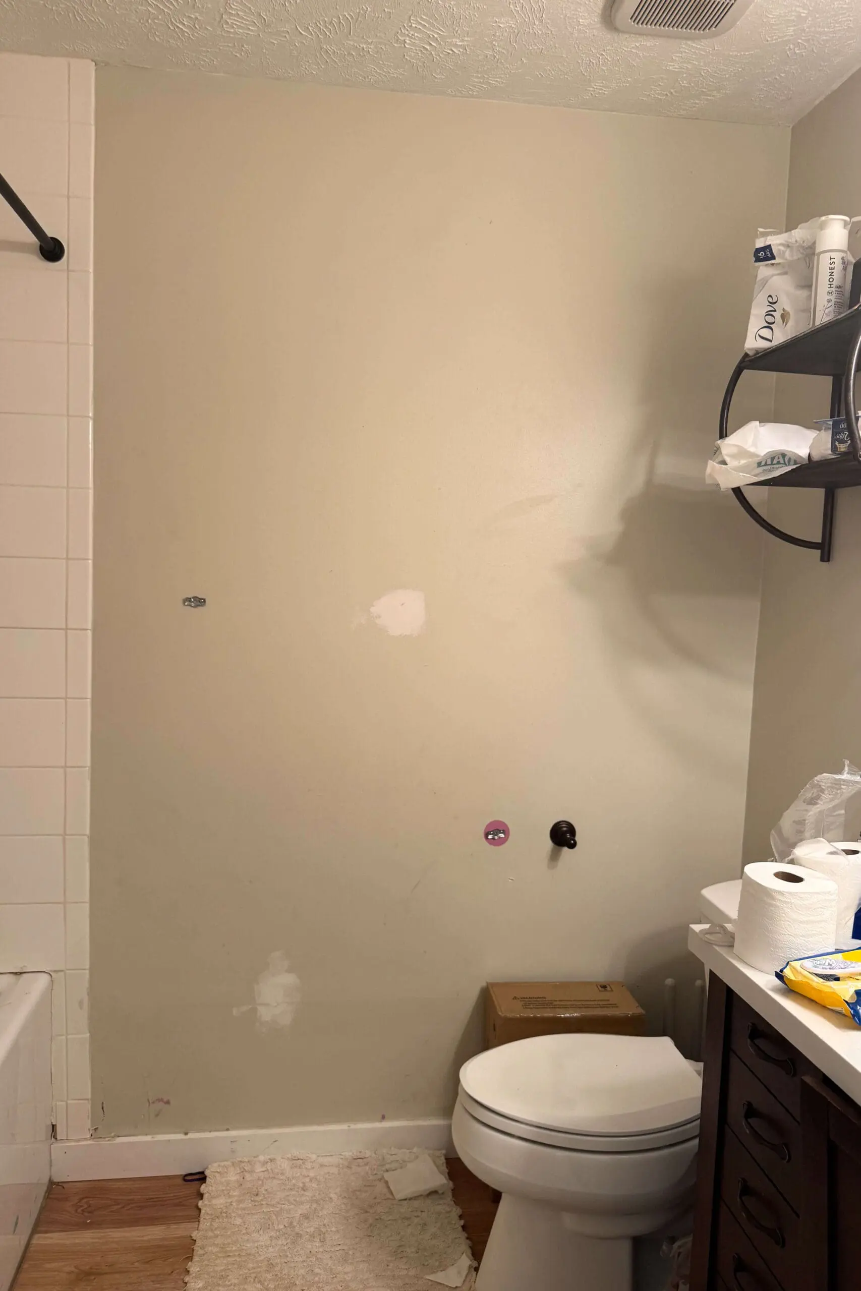 A beige bathroom wall with visible patches from previous repairs. Below, a standard white toilet sits near a small wooden cabinet with drawers. An open black metal shelf above holds toilet paper rolls and personal care items. The flooring appears to be laminate or vinyl with a light wood pattern.