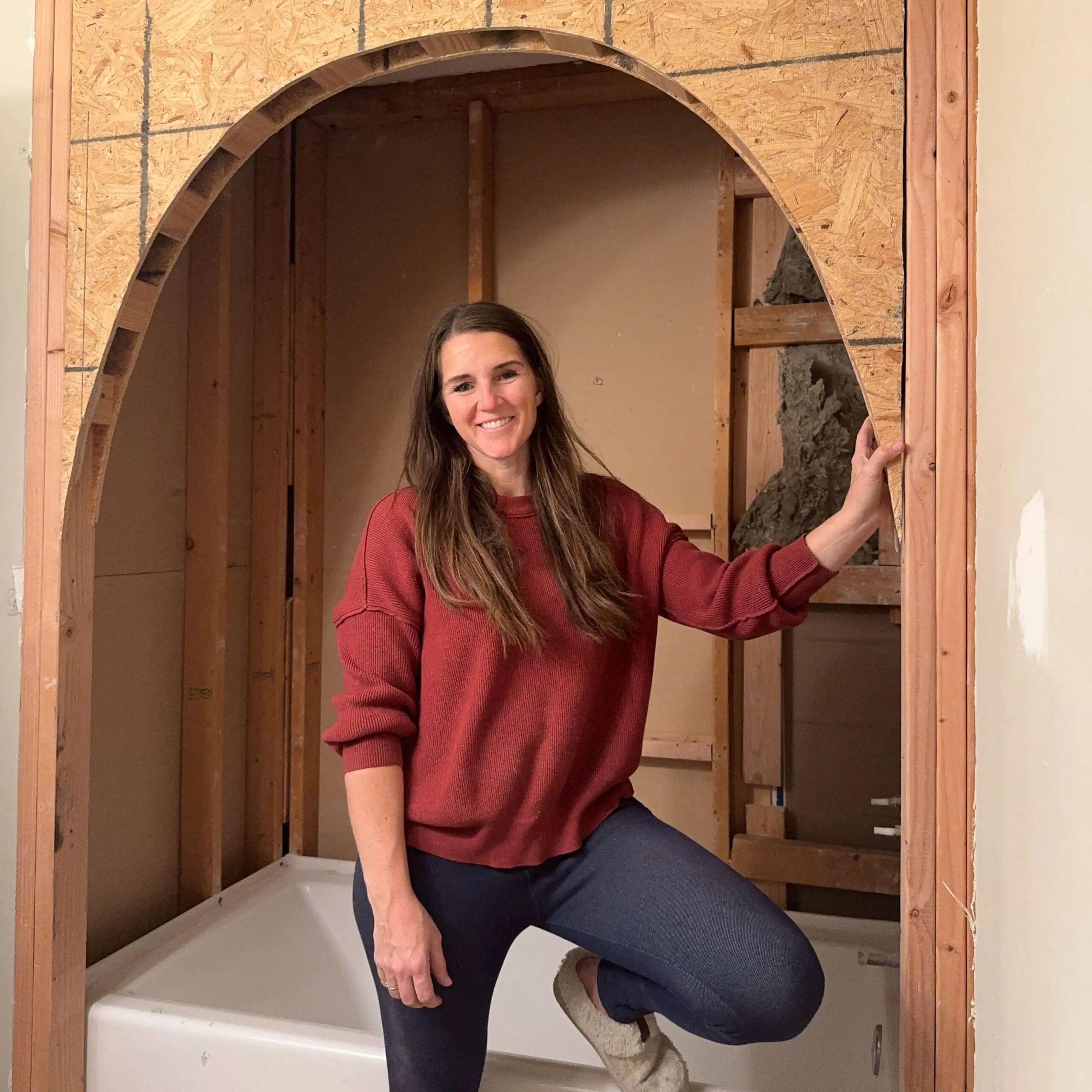 A woman in a red sweater and blue jeans stands inside a bathroom remodel next to an arched bathtub alcove she constructed. The exposed studs and OSB board create a dramatic, custom architectural feature over the bathtub.