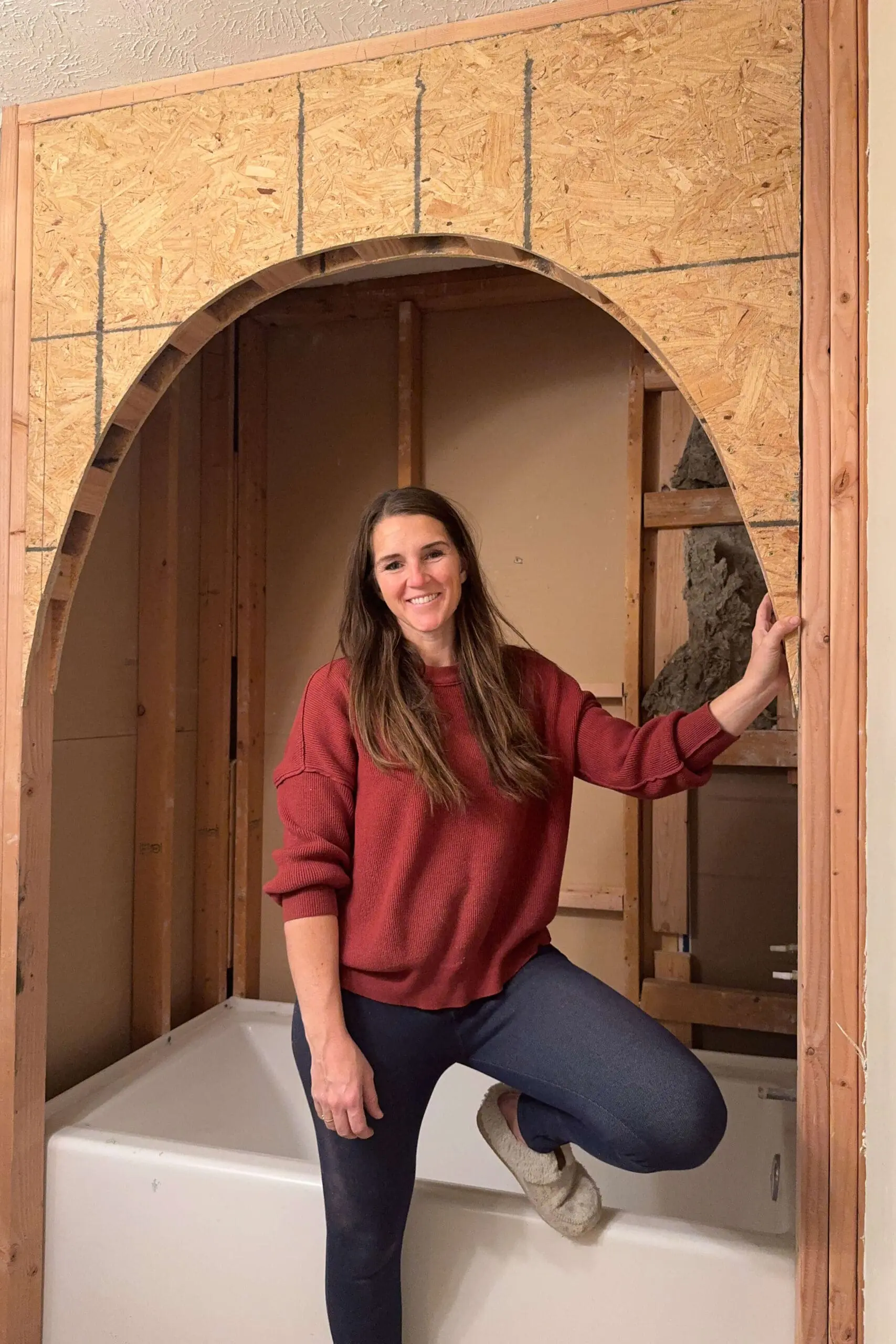 A woman in a red sweater and blue jeans stands inside a bathroom remodel next to an arched bathtub alcove she constructed. The exposed studs and OSB board create a dramatic, custom architectural feature over the bathtub.