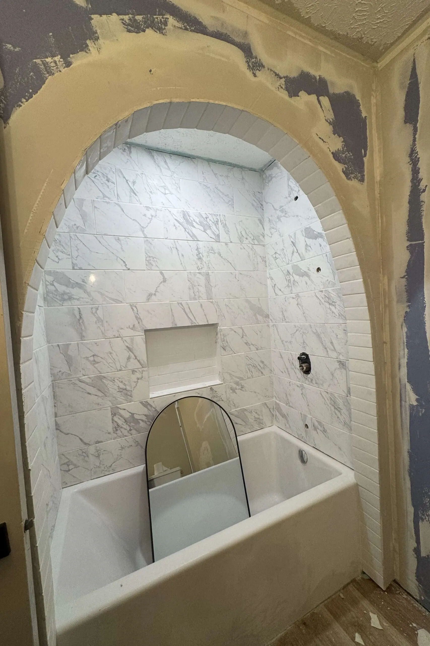 A completed bathroom shower with an arched opening, tiled with marble-style tiles. The recessed niche is tiled with white subway tiles, and the curved arch is finished with small white tiles. A mirror leans inside the bathtub beneath the shower.