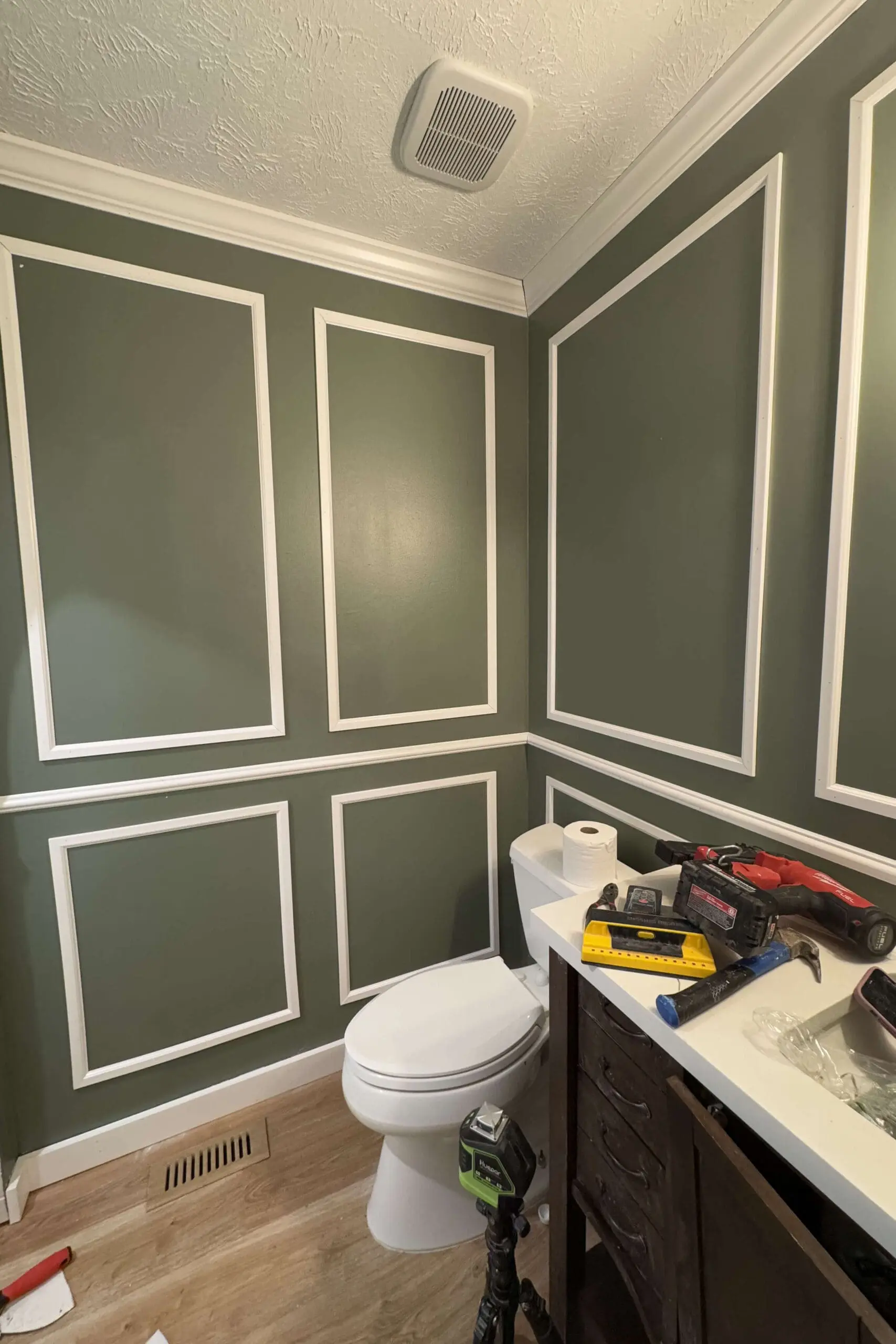 Bathroom makeover in progress featuring a green wall with DIY picture frame molding, a white toilet, and tools scattered on the vanity, illustrating a DIY project setup.