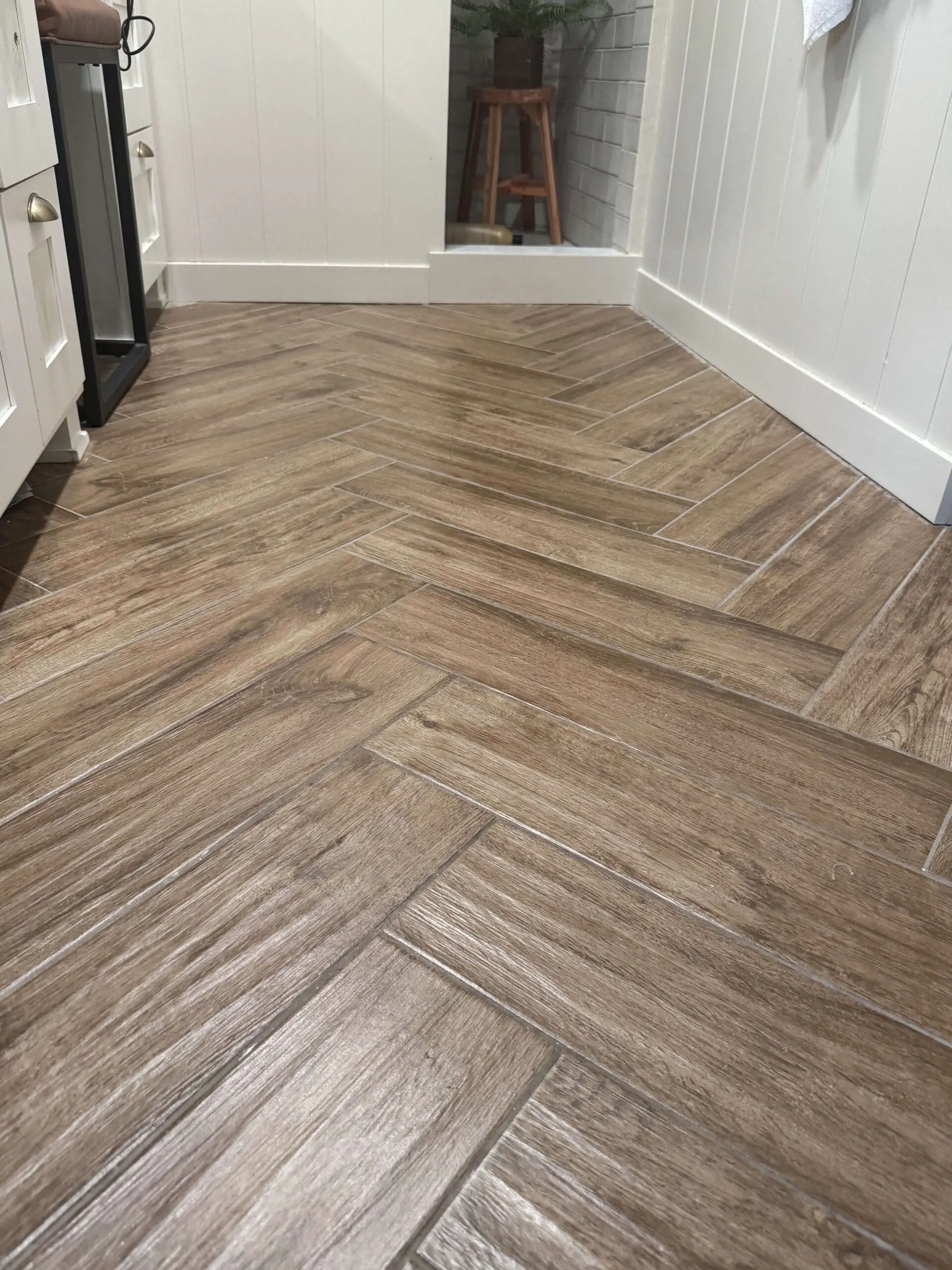 A closer view of a bathroom floor showcasing herringbone-patterned wood-look tiles with subtle texture, surrounded by crisp white baseboards, leading to a shower area with white subway tiles and a wooden stool for decor.