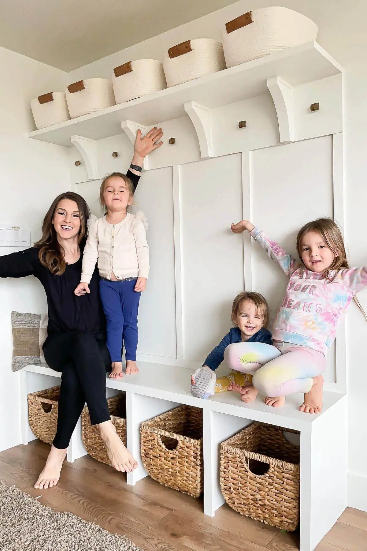 A smiling woman and three young children sitting and playing on a completed built-in entryway bench with storage cubbies and a white board and batten wall. The finished DIY project features woven baskets, decorative hooks, and a top shelf with neutral fabric bins, creating a stylish and functional mudroom space.