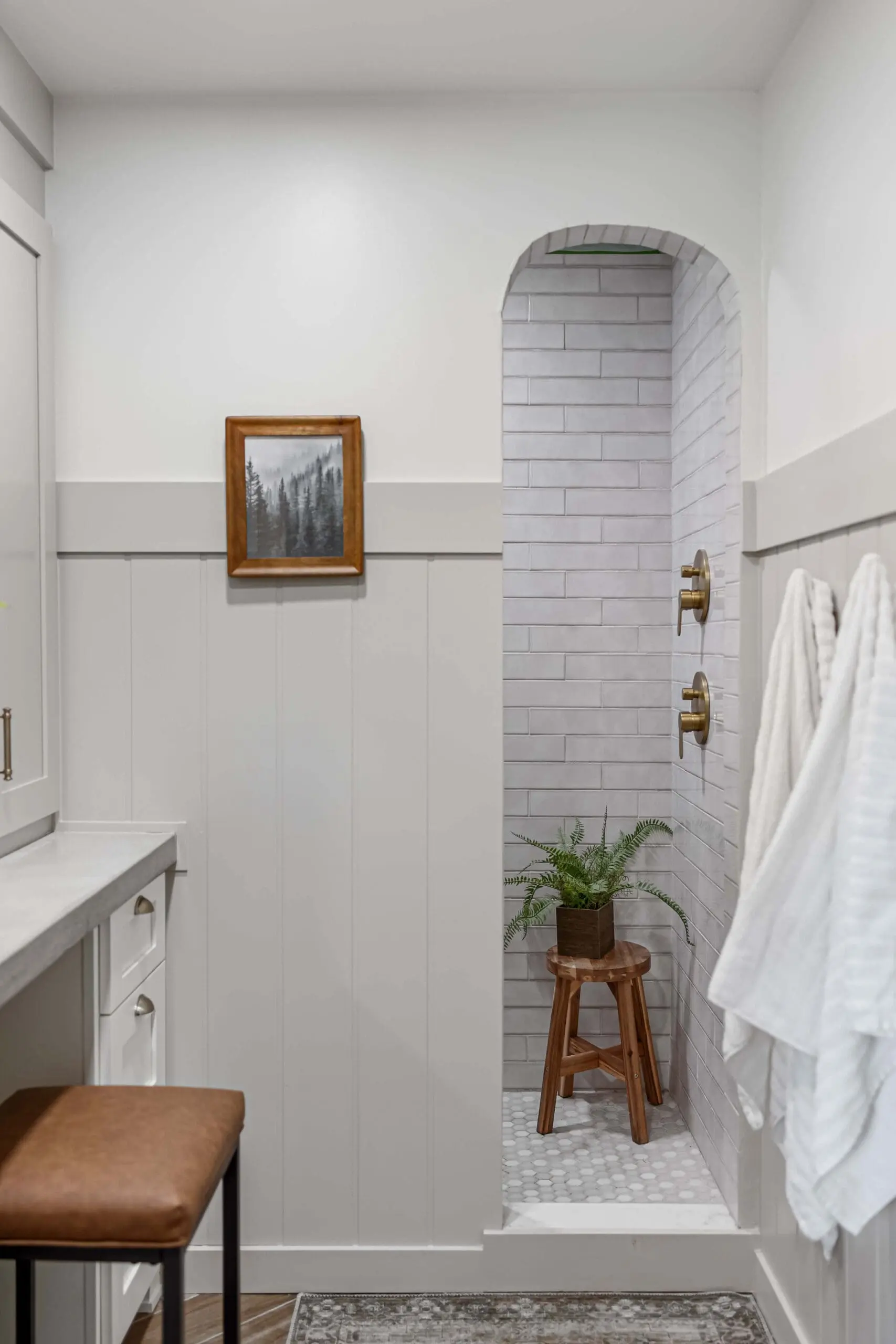 A warm and inviting bathroom showcasing a minimalist arched shower entrance with elegant gray subway tiles and brass shower fixtures. The neutral color palette is enhanced by soft white board and batten walls, a wooden vanity stool with a tan leather cushion, and natural décor like woven baskets and wooden shelving.