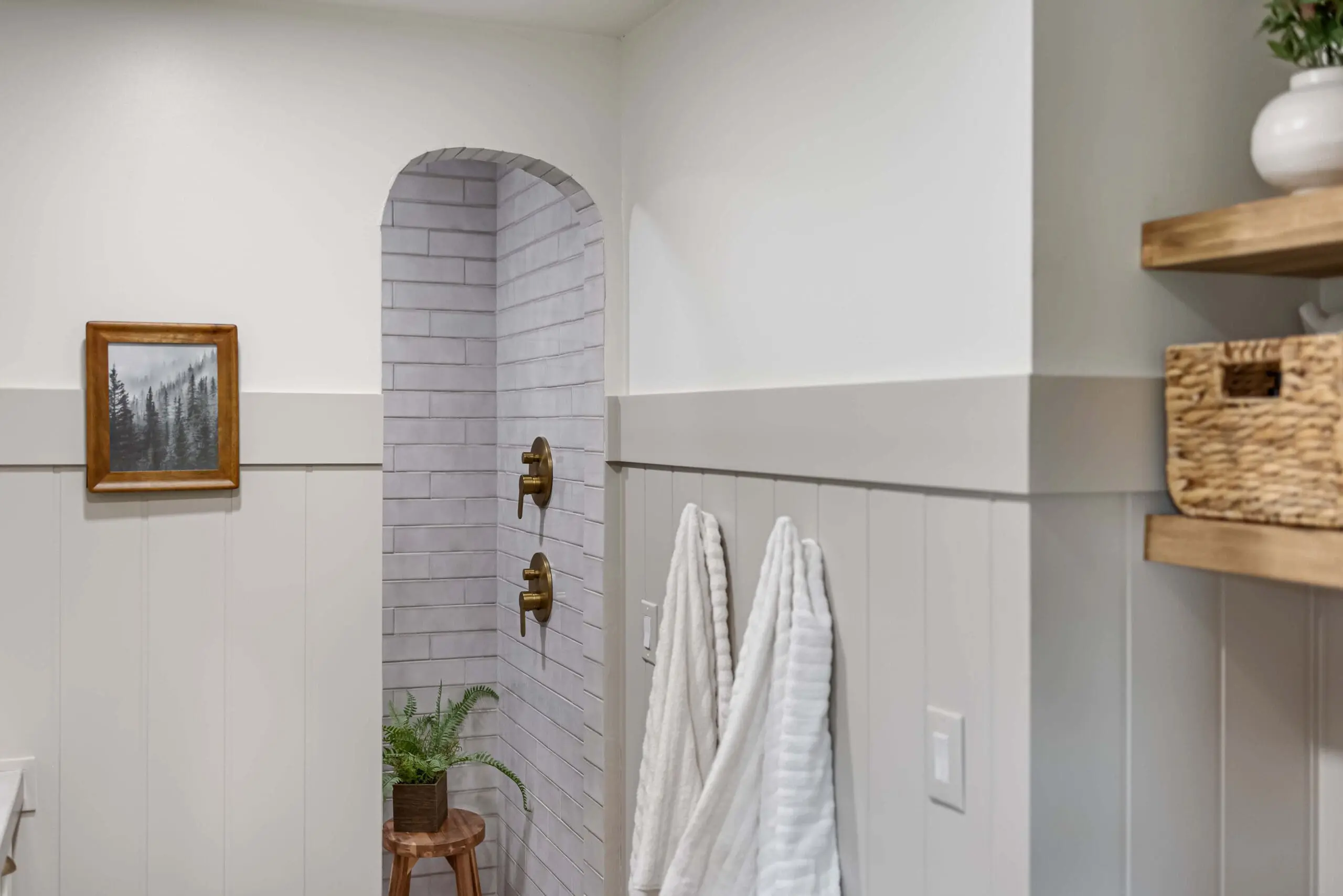 A modern farmhouse-style bathroom with a serene arched walk-in shower, lined with light gray subway tiles and dual brass shower controls. The space is accented with a natural wood stool, a green plant, and soft textured towels hanging on the wall. White board and batten paneling adds a cozy yet sophisticated touch.