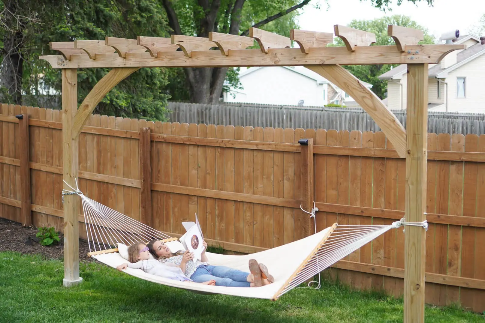 A beautiful wooden pergola with a built-in hammock stands in a lush backyard, supported by sturdy posts with decorative curved beams. Two children relax in the hammock, reading a book together, creating a peaceful outdoor retreat. A wooden privacy fence encloses the yard, with greenery and trees in the background.