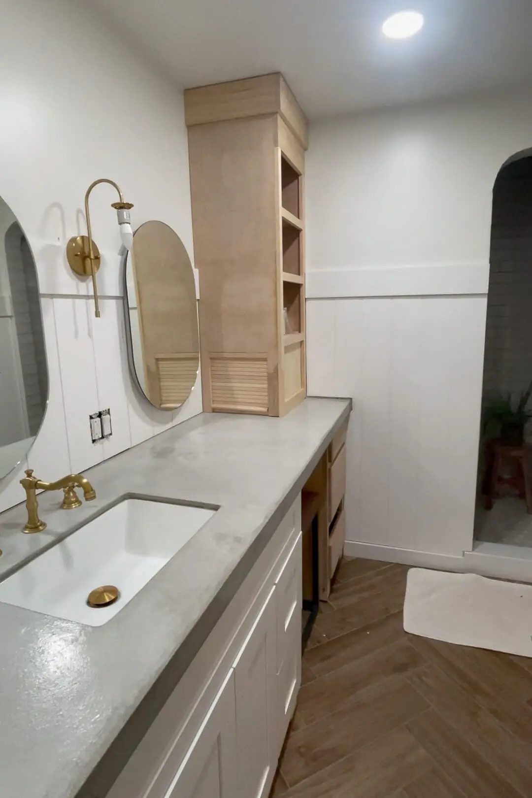 Primary bathroom remodel in progress featuring a modern farmhouse vanity with a sleek concrete countertop, brass faucet, and white shaker-style cabinetry. A custom-built wooden storage tower is partially installed, adding functionality to the space. The round, frameless mirror is complemented by a gold wall sconce, while white vertical paneling adds texture. The warm wood-look tile flooring contrasts beautifully with the light and airy design.