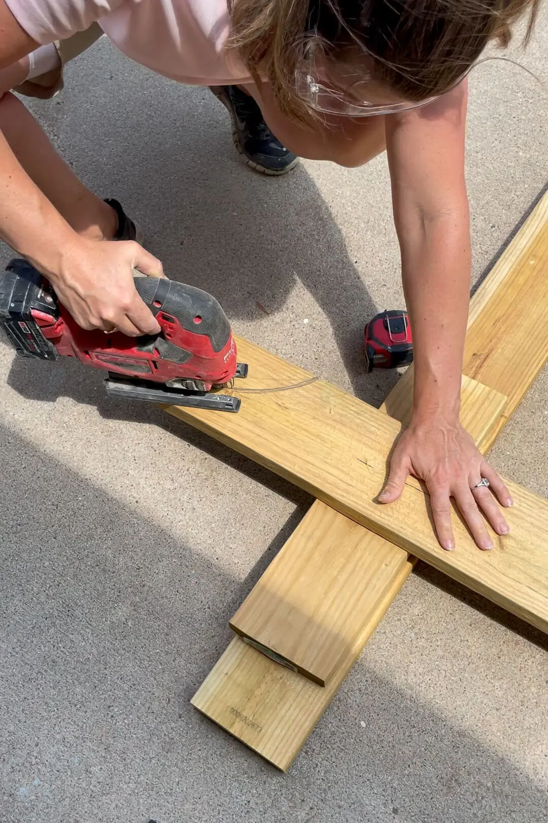 A woman wearing safety glasses and a pink shirt uses a red jigsaw to cut a curved design into a wooden board. She firmly holds the board in place while cutting, with a red tape measure beside her on the concrete.
