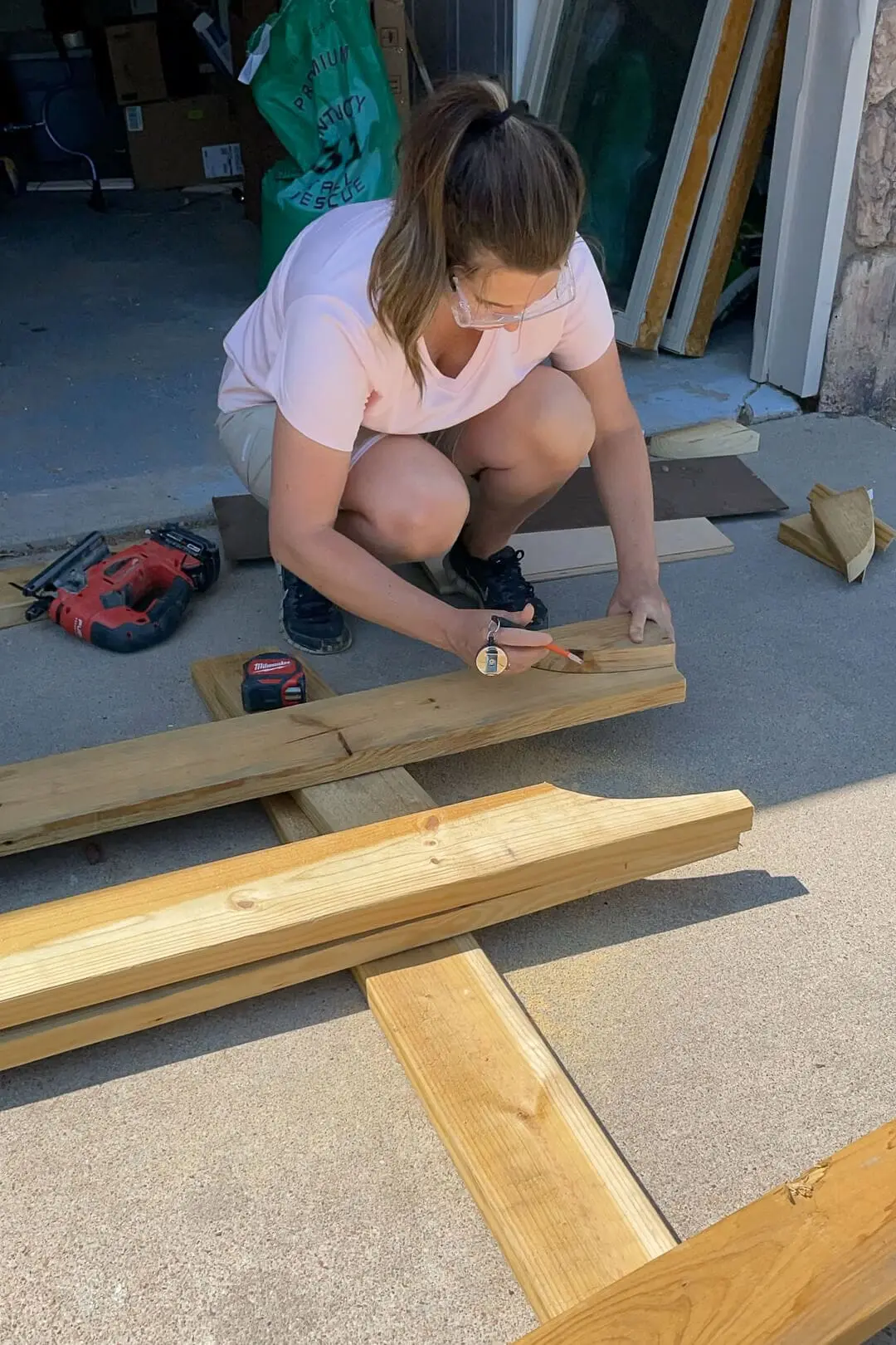 A woman in a pink shirt and safety glasses crouches over a wooden beam, carefully marking a design for a garden arbor with a pencil. Various woodworking tools, including a jigsaw and a measuring tape, are scattered around her outdoor workspace near a garage.