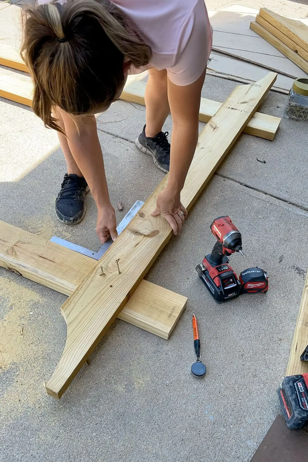A woman positions a wooden beam while using a speed square to ensure alignment. A red power drill, tape measure, and other woodworking tools are nearby as she prepares to secure the beam in place.