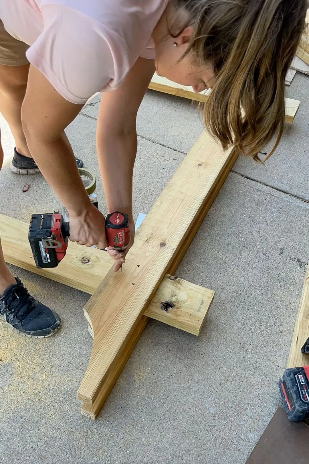 A woman in a pink shirt and black sneakers drills a screw into a wooden beam, securing a decorative cut piece. Sawdust and various woodworking tools are scattered on the concrete as she constructs an outdoor project.