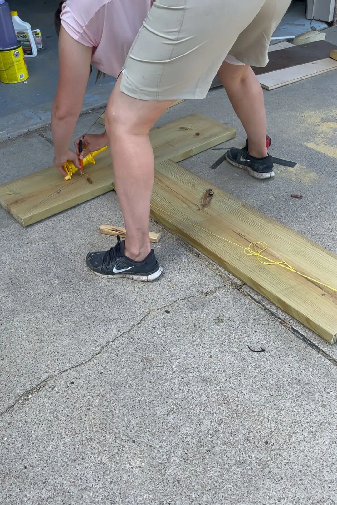 A woman in beige shorts and a pink shirt bends over a wooden structure on a concrete driveway, using a carpenter's string to mark precise measurements. The surrounding area has woodworking tools, sawdust, and cut lumber.