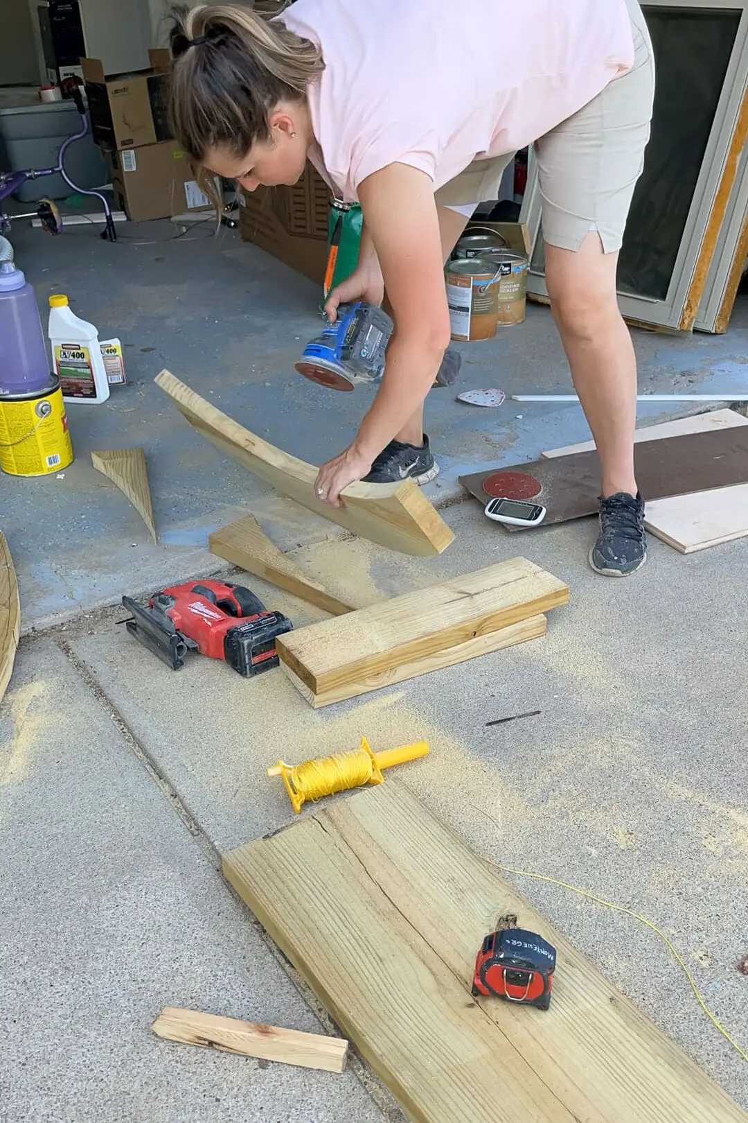 A woman in a pink shirt sands a curved wooden piece with a power sander, smoothing out the edges for a woodworking project. Various tools, sawdust, and materials are scattered in an open garage workspace.