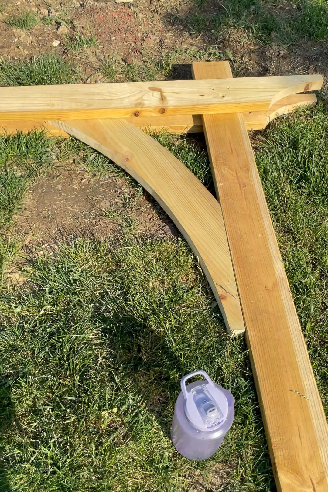 A partially assembled wooden garden arbor structure lays on the grass, with a large water bottle beside it. The curved beam is securely attached, showing progress in the DIY project.

