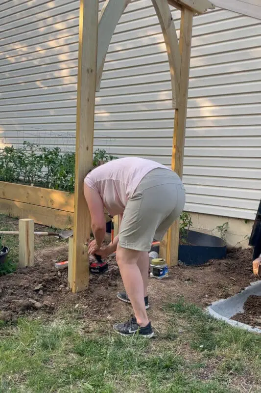 A woman wearing a pink shirt and beige shorts bends over, using a power drill to secure a wooden post into the ground as part of a DIY garden arbor project.