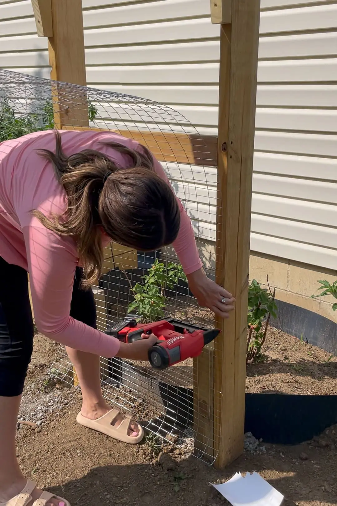 A woman in a pink long-sleeve shirt uses a red nail gun to attach wire mesh to a wooden garden arbor. The structure is partially completed, with plants growing inside the enclosed area.