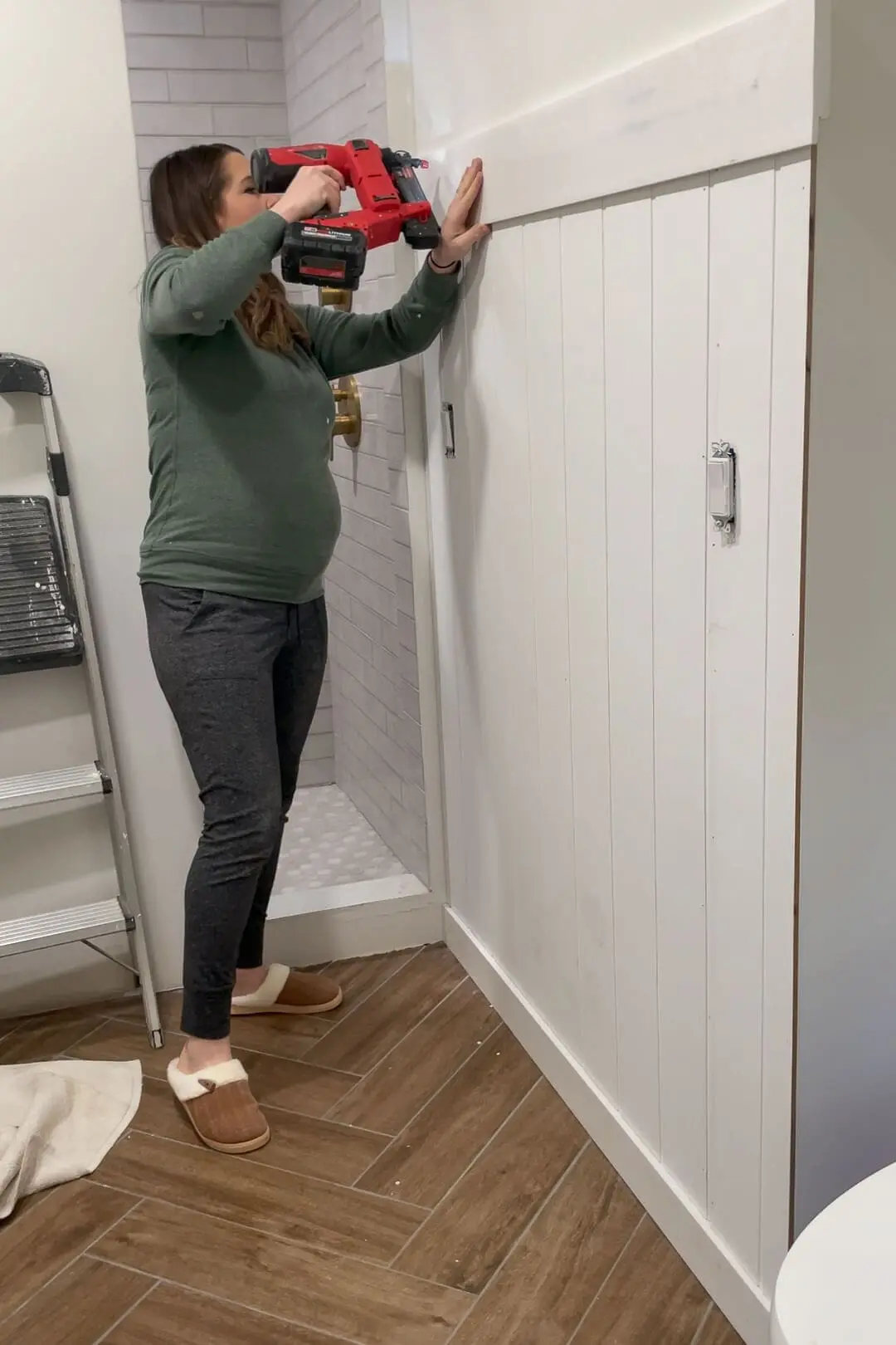 A woman standing in a bathroom, securing a horizontal trim board at the top of a newly installed vertical shiplap wall using a red nail gun. The DIY renovation highlights a fresh and modern design, featuring brass shower fixtures and white subway tiles in an arched shower alcove.