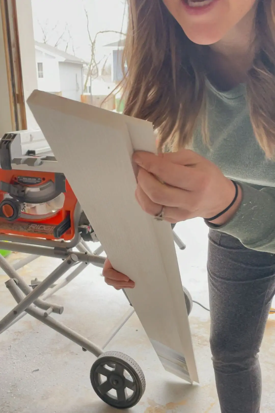 A close-up shot of a woman holding a cut piece of white pvc trim with a custom groove cut for capping the top of vertical shiplap walls. The background reveals a woodworking setup with a Ridgid table saw in a well-organized garage workspace.