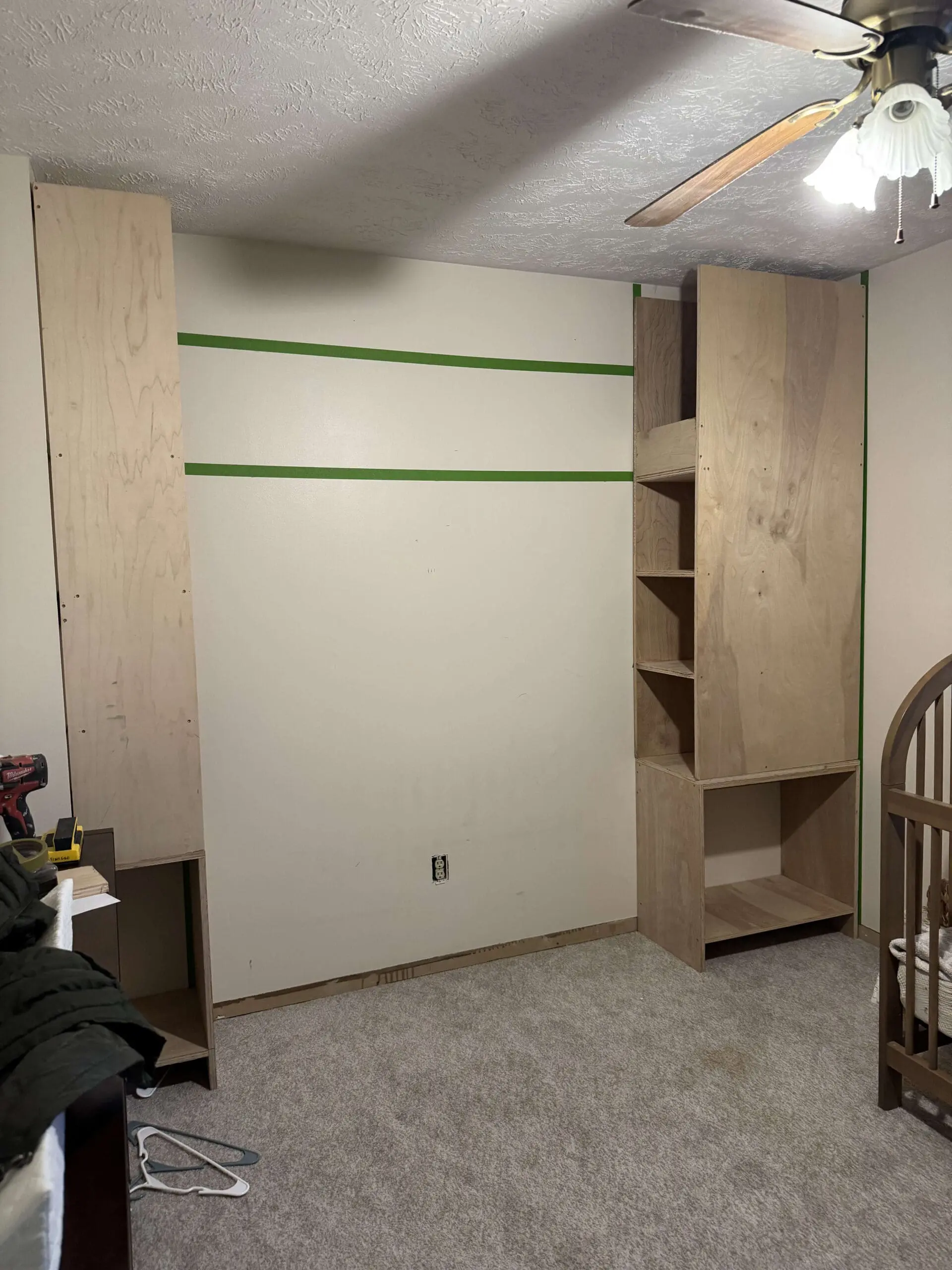 A partially completed DIY built-in shelving unit in a bedroom. Two tall wooden storage units made of plywood are installed on either side of a beige wall, with green painter’s tape marking future design elements. The left unit is an open vertical panel, while the right unit has multiple shelves and a lower open compartment. A crib is partially visible in the foreground, and a ceiling fan with a light fixture hangs above. Tools and materials are scattered nearby, showing an active construction project in progress.