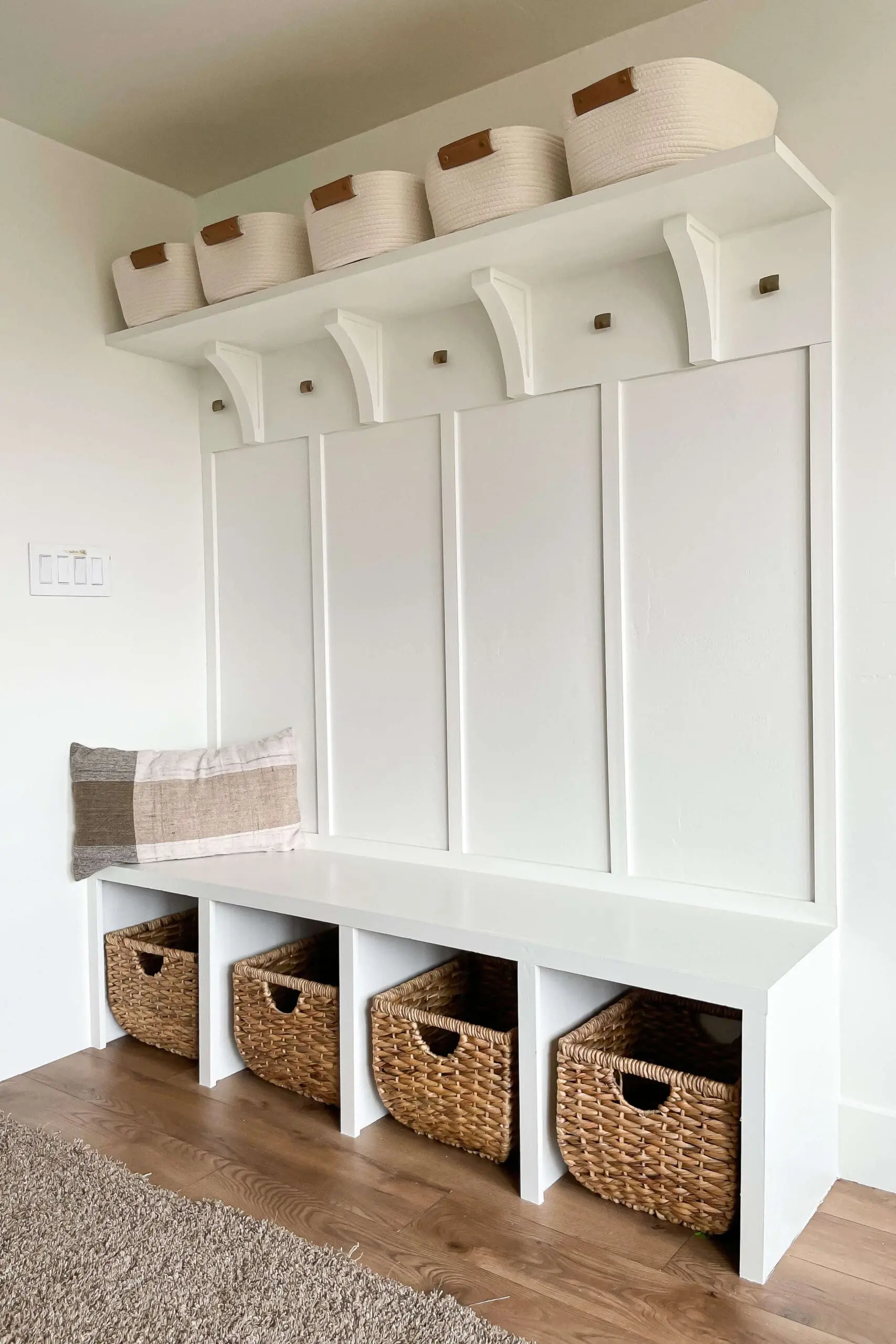 Stylish and functional mudroom storage setup with a white built-in bench, four cubbies with woven baskets, a board and batten wall with wooden hooks, and a floating shelf holding neutral fabric baskets.