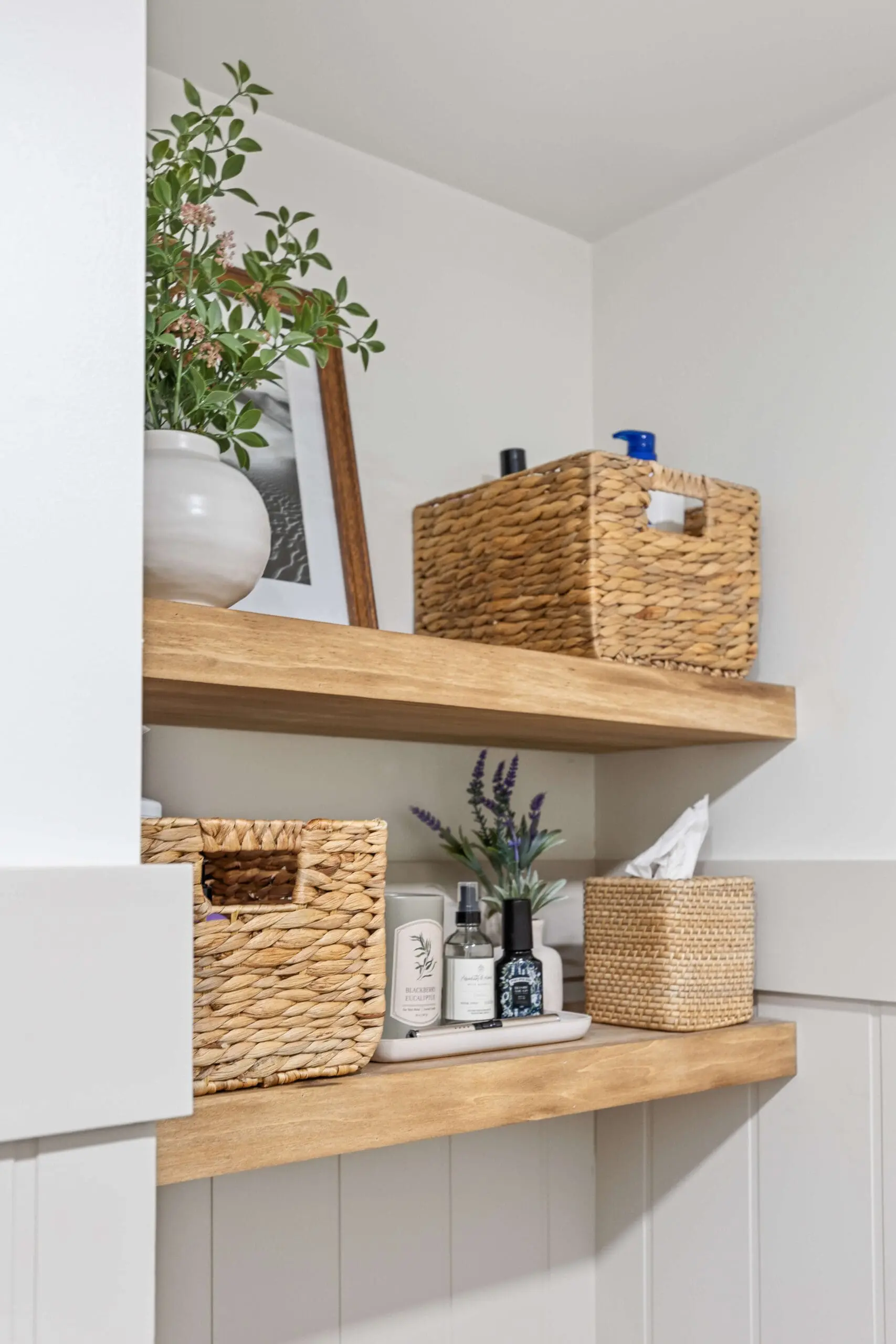 Two wooden floating shelves in a bathroom corner, styled with woven baskets, glass bottles, and a potted plant in a white ceramic vase. A framed black and white photograph leans against the wall, adding a personal touch. The natural textures contrast beautifully with the white paneled walls, creating a warm and organized space.