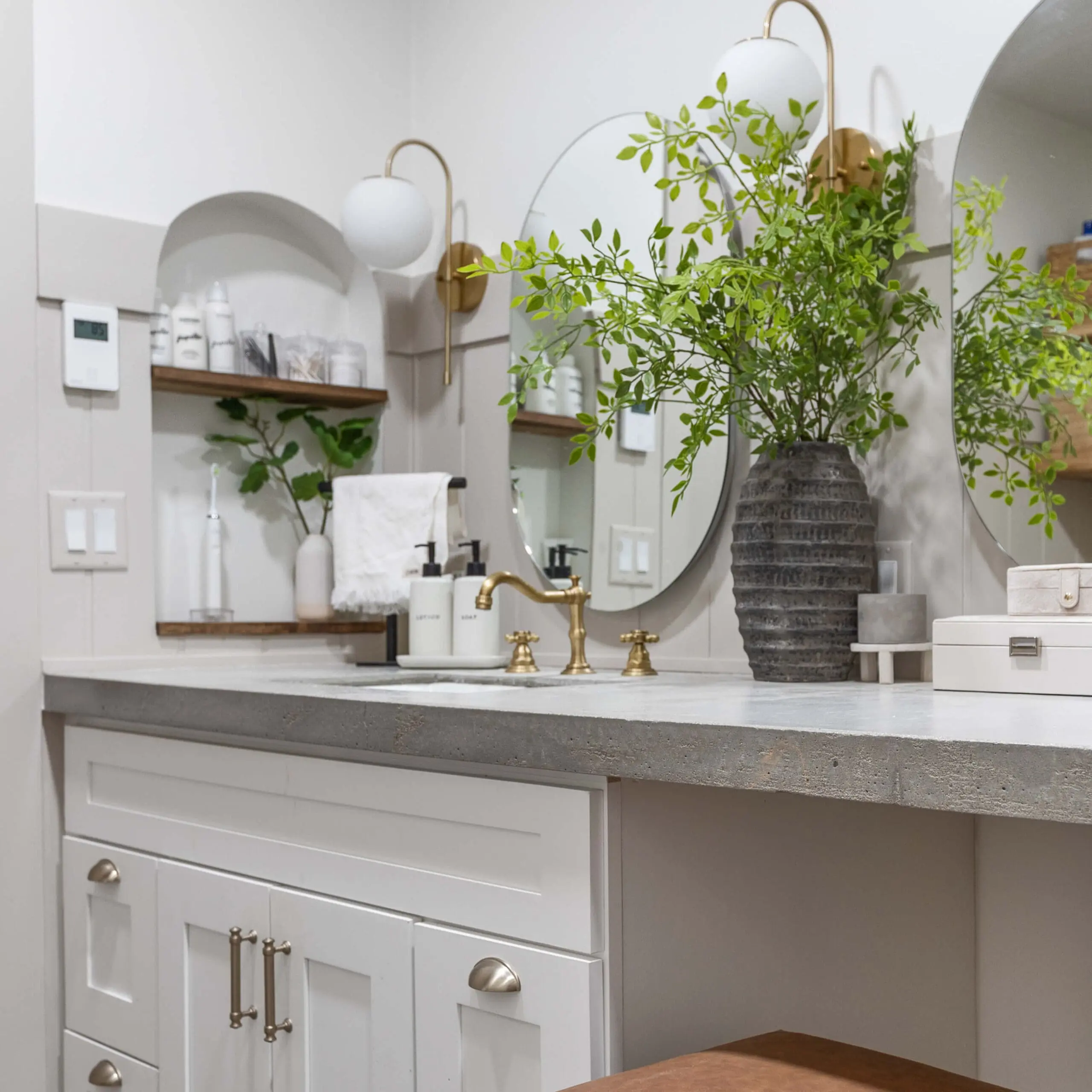 Modern farmhouse bathroom with a sleek concrete countertop, brass fixtures, and white shaker-style cabinetry. A large, round mirror is flanked by elegant gold and glass sconces, reflecting the fresh greenery in a textured black vase. Built-in arched shelves hold neatly organized toiletries, adding a touch of functionality and charm to the serene, neutral-toned space. This bathroom is the after of a DIY primary bath remodel.