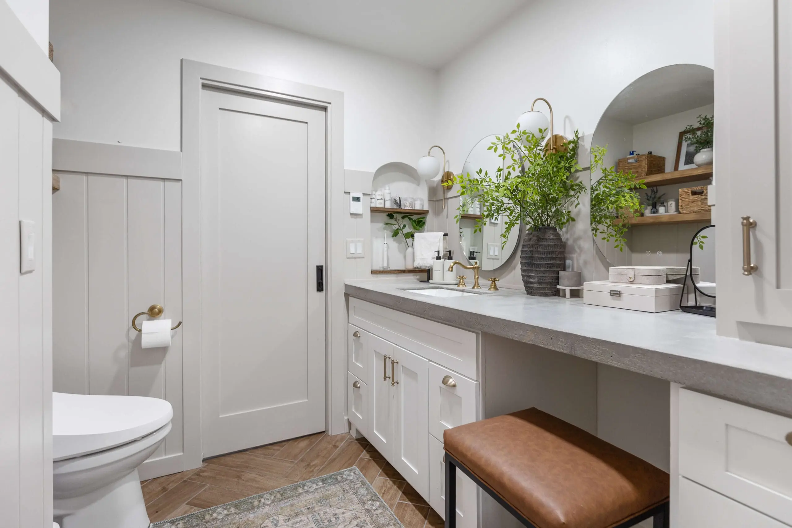 Primary bath remodel with a sleek, modern farmhouse vanity featuring a concrete countertop, brass fixtures, and white shaker cabinets. A soft neutral sliding door with a matte black handle leads to an adjoining space. The floor is finished in a warm wood-look tile, adding texture and contrast.