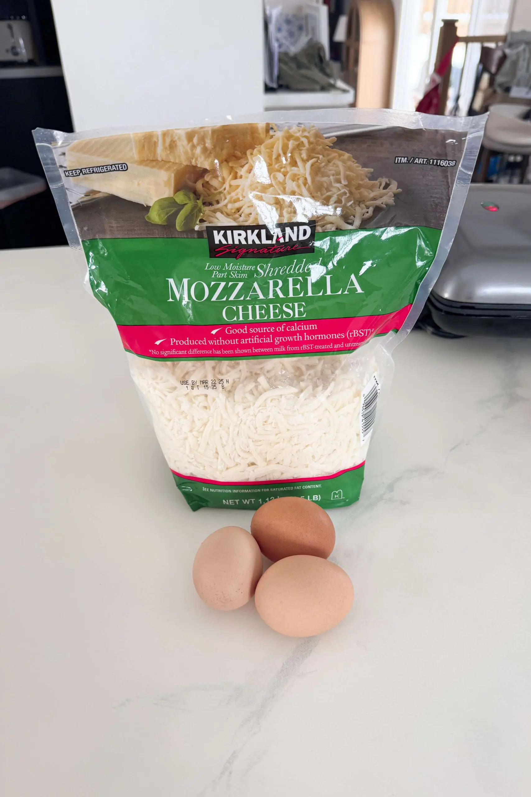A bag of Kirkland Signature shredded mozzarella cheese sits on a white kitchen counter, with three fresh brown eggs placed in front, ready to be made into a carnivore diet breakfast. A waffle maker and kitchen essentials are visible in the background.
