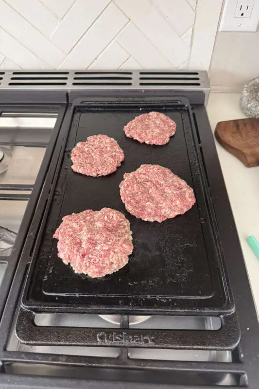 Four raw sausage patties sizzling on a black stovetop griddle, ready to be cooked. The kitchen background features a white herringbone tile backsplash and countertop essentials.