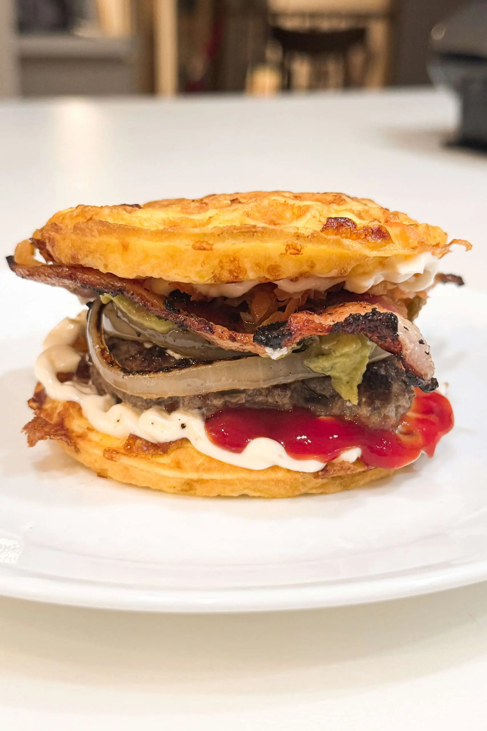 A close-up of a carnivore bread burger with crispy bacon, caramelized onions, and a thick beef patty, all sandwiched between two golden, cheese-infused chaffle (cheese waffle) buns. The burger is layered with a generous amount of mayo and ketchup, with some sauce slightly oozing out.