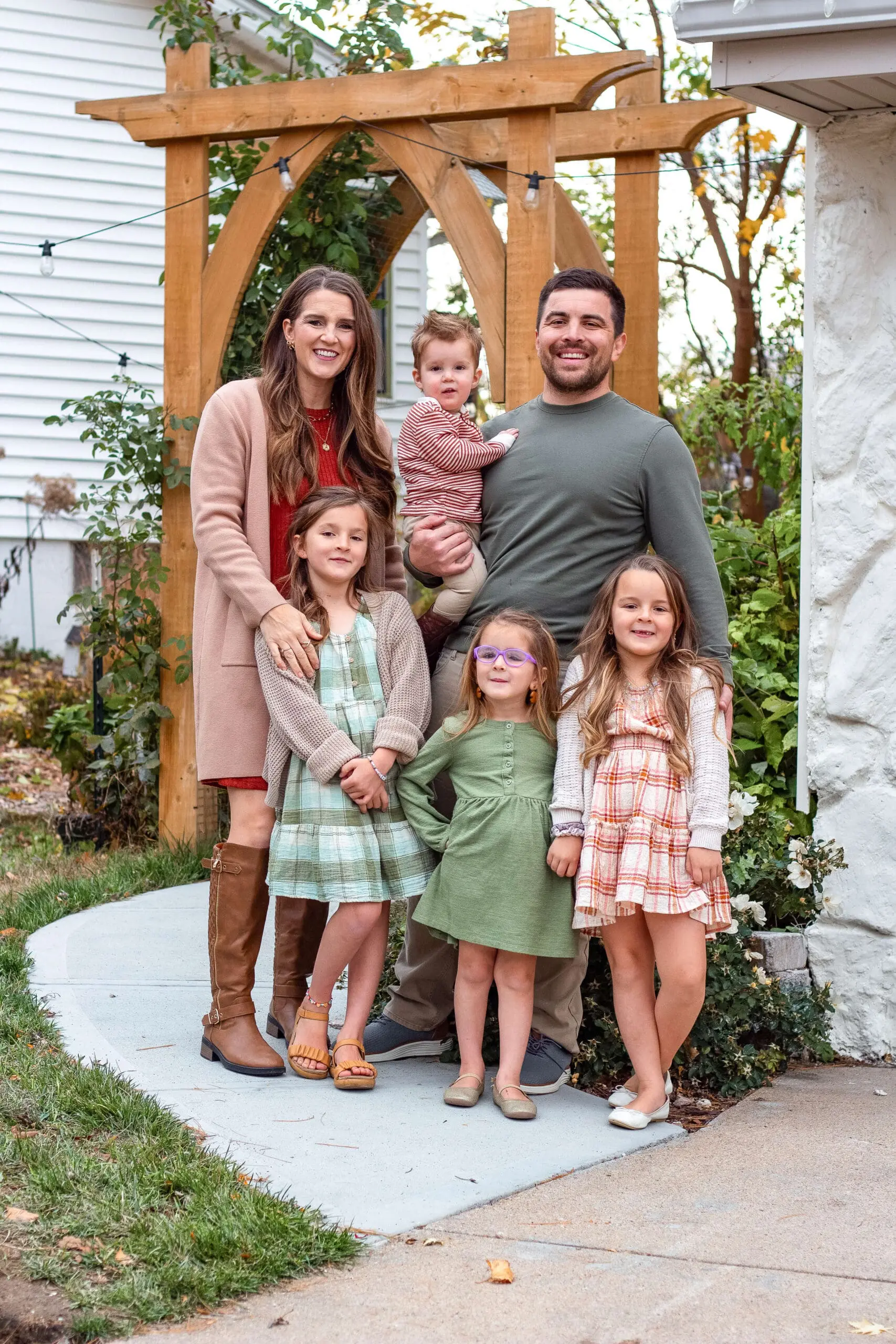 A family of six poses for a portrait in front of a handcrafted wooden garden arbor, surrounded by lush greenery. They are dressed in coordinating fall outfits, with warm tones and soft pastels, creating a cozy and welcoming scene.
