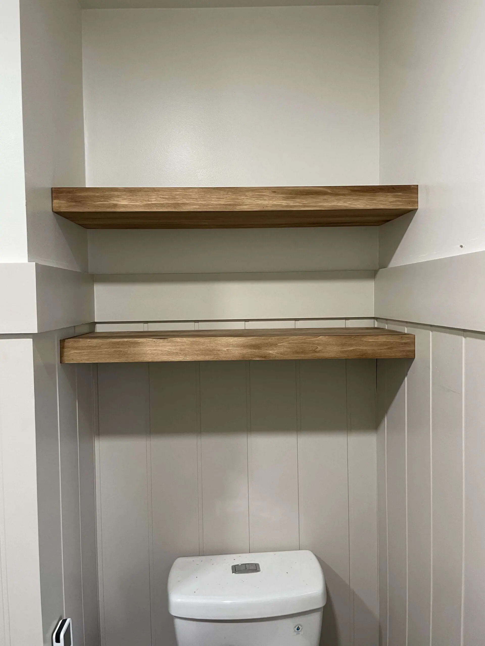 Primary bath remodel featuring newly installed wooden floating shelves above the toilet. The warm wood finish contrasts with the light, neutral paneling, adding functional storage space. The remodel is near completion, with clean lines and a fresh, modern farmhouse aesthetic.