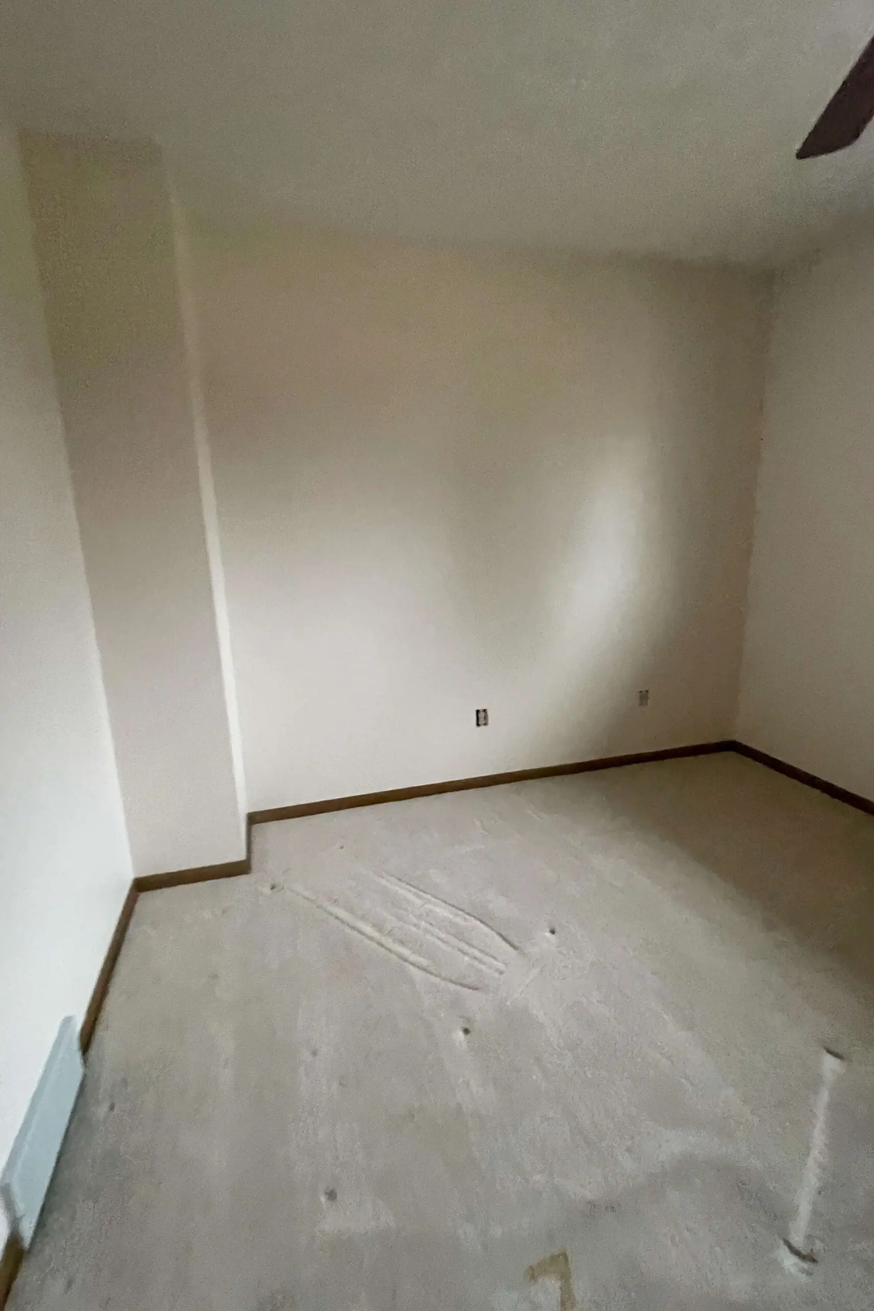 An empty bedroom with freshly cleaned beige walls and light-colored carpet, showing faint impressions from previously placed furniture. The room has a simple layout with a white air vent near the floor and electrical outlets on the wall. The space is now cleared out, ready for a transformation.