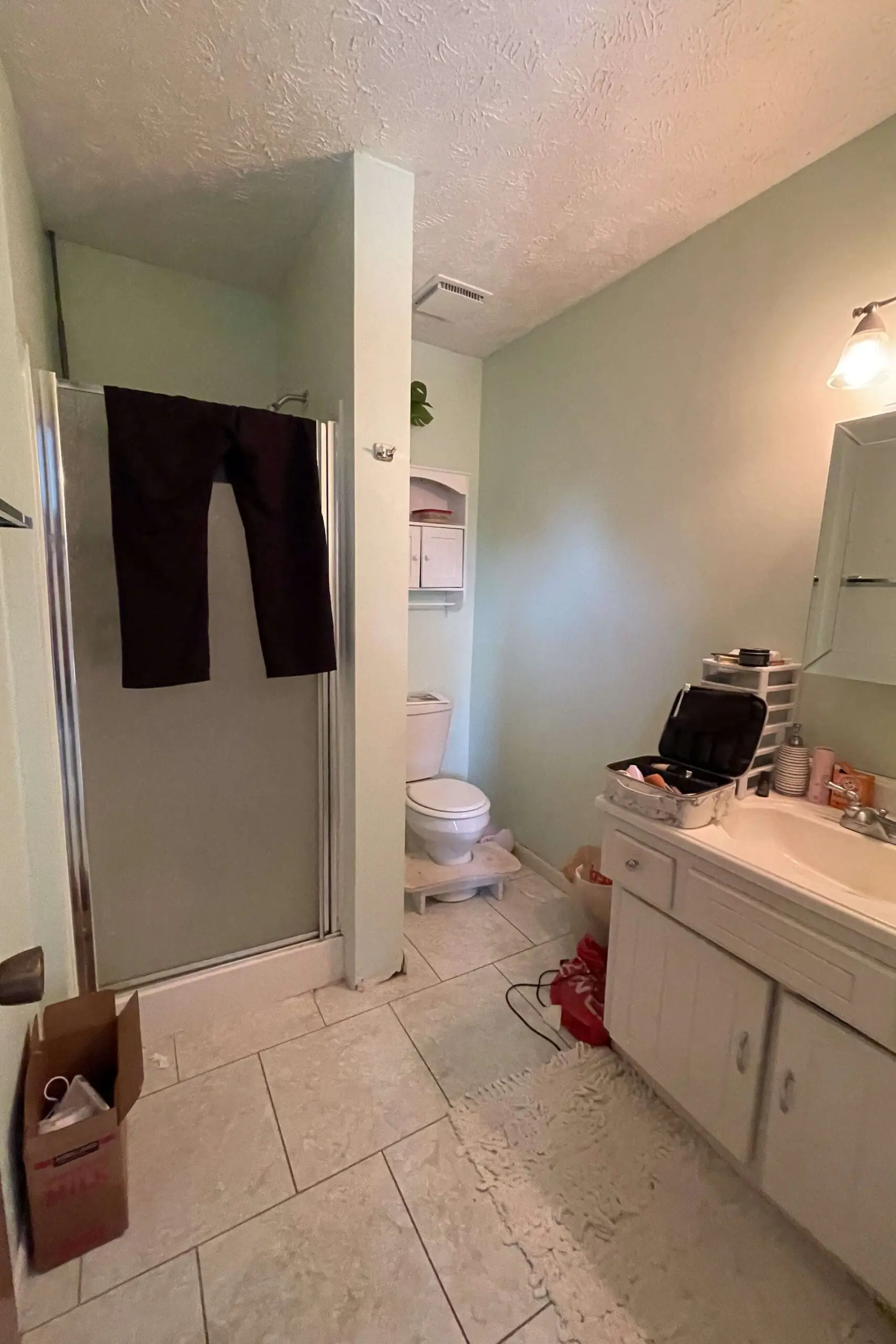Outdated bathroom about to undergo a primary bath remodel with pale green walls, a compact shower with a sliding door, and a basic white vanity. A pair of dark pants hangs over the shower door, and various toiletries clutter the countertop. The space has dated tile flooring, and the toilet area features a small built-in cabinet