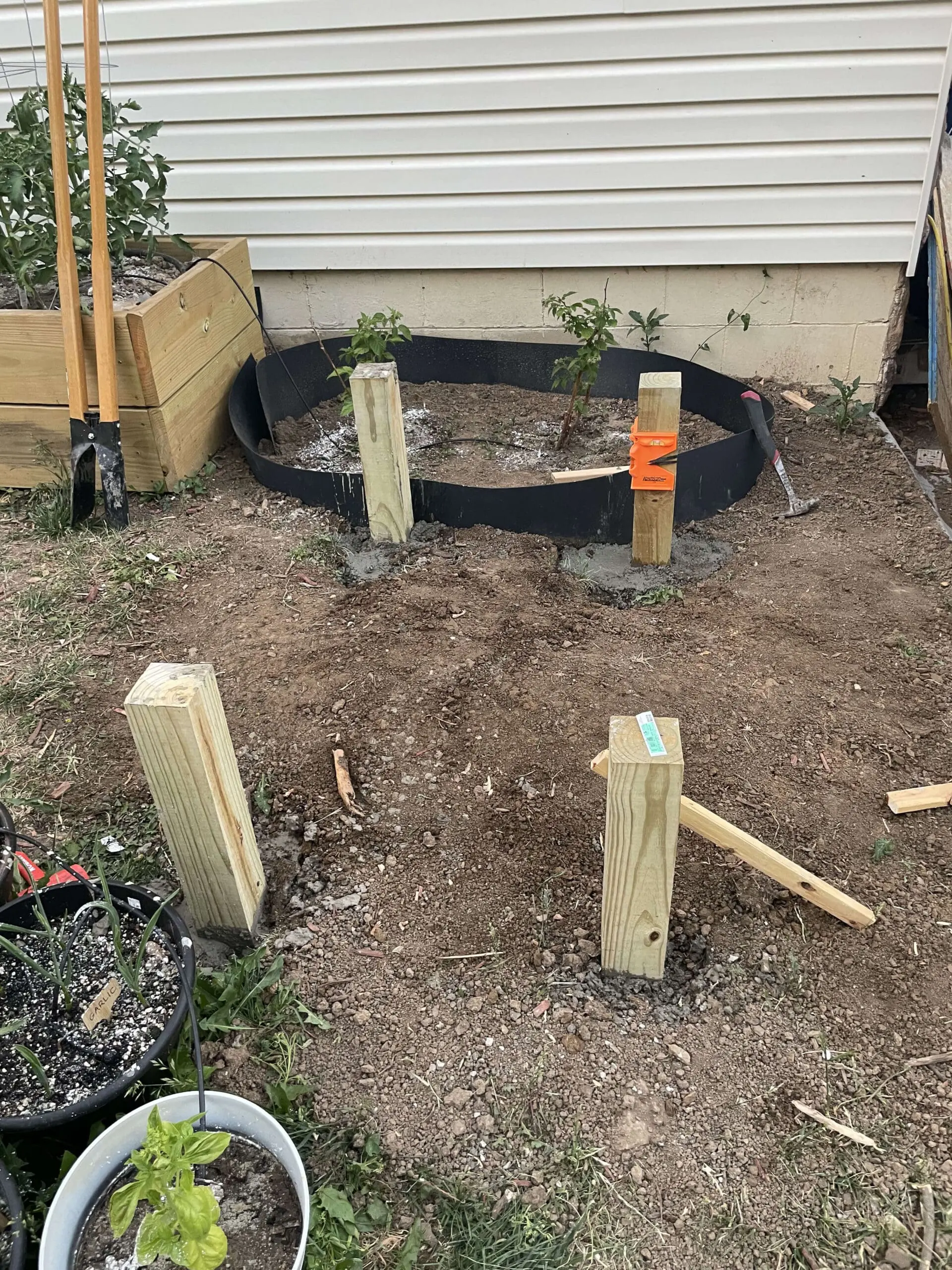 Four wooden posts are set in concrete, marking the foundation of a garden arbor. A black landscape border surrounds a small garden bed, and various tools and a post hole digger are nearby.